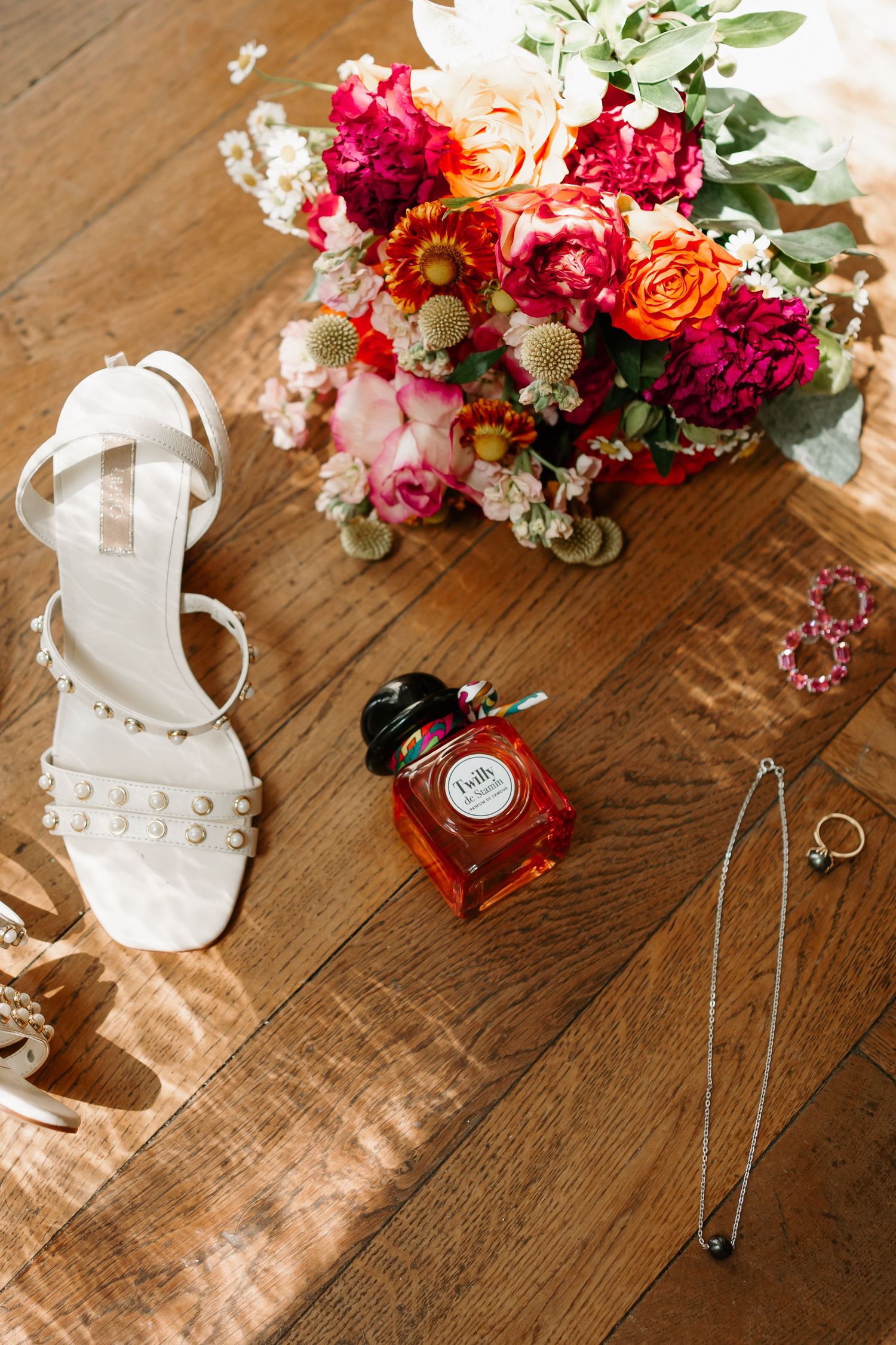 White bridal sandals, floral bouquet with pink and orange blooms, and wedding accessories arranged on wooden floor.