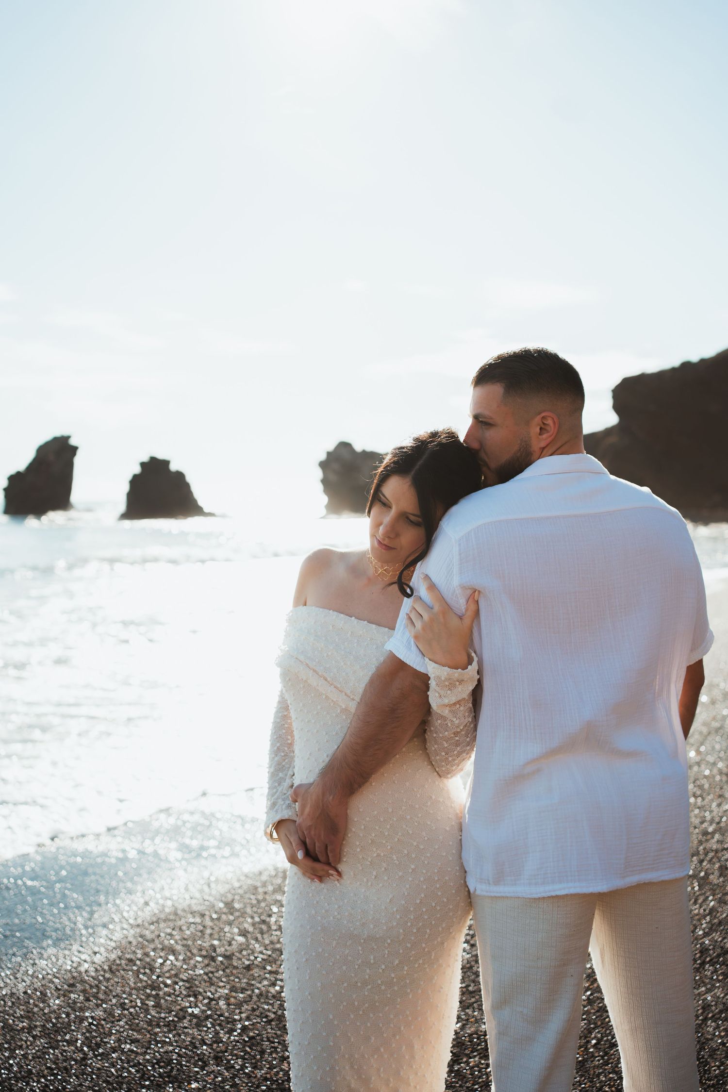 Couple in white embraces on rocky beach shore during sunset.