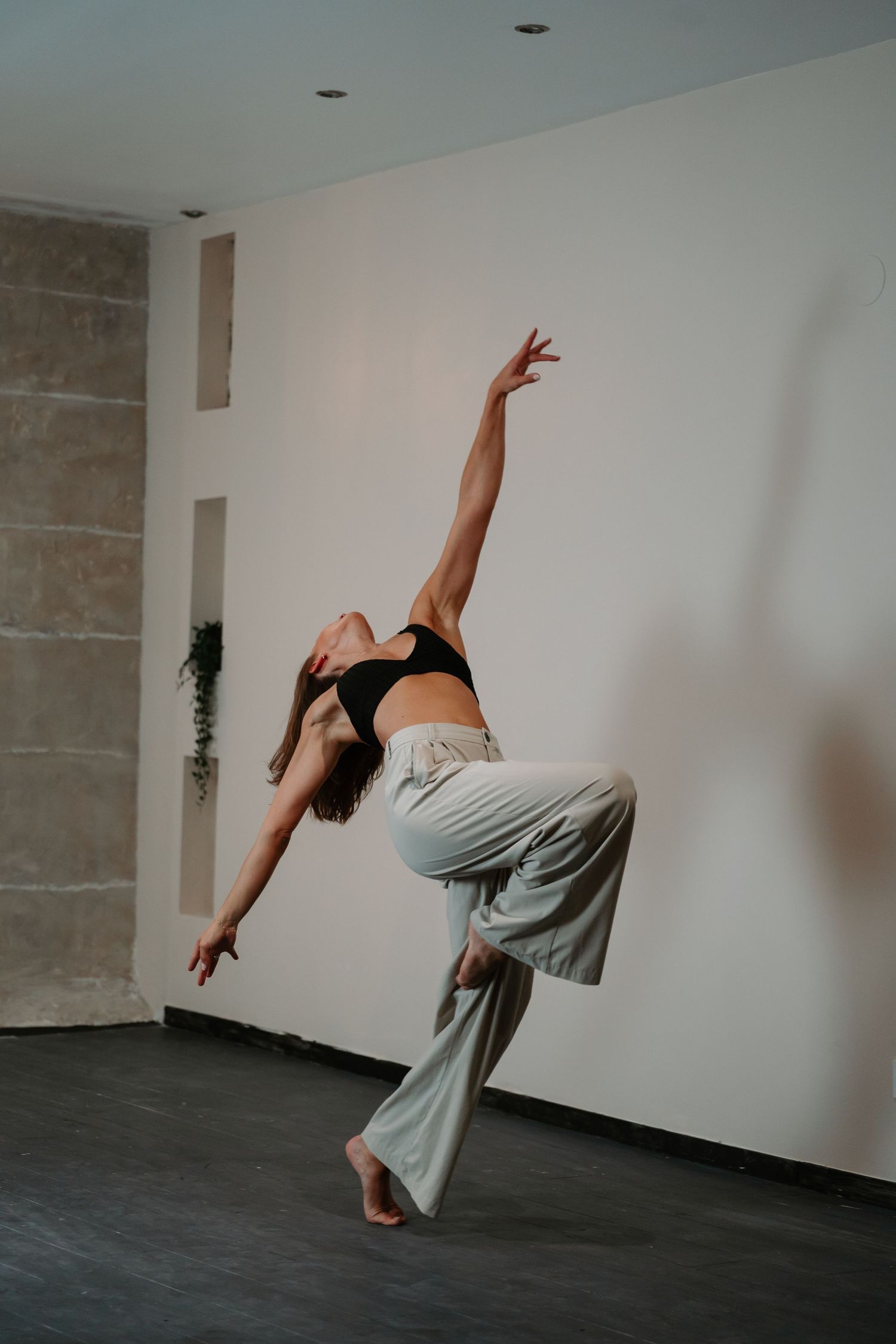 Contemporary dancer in black crop top and flowy gray pants performs an expressive movement against white studio wall.