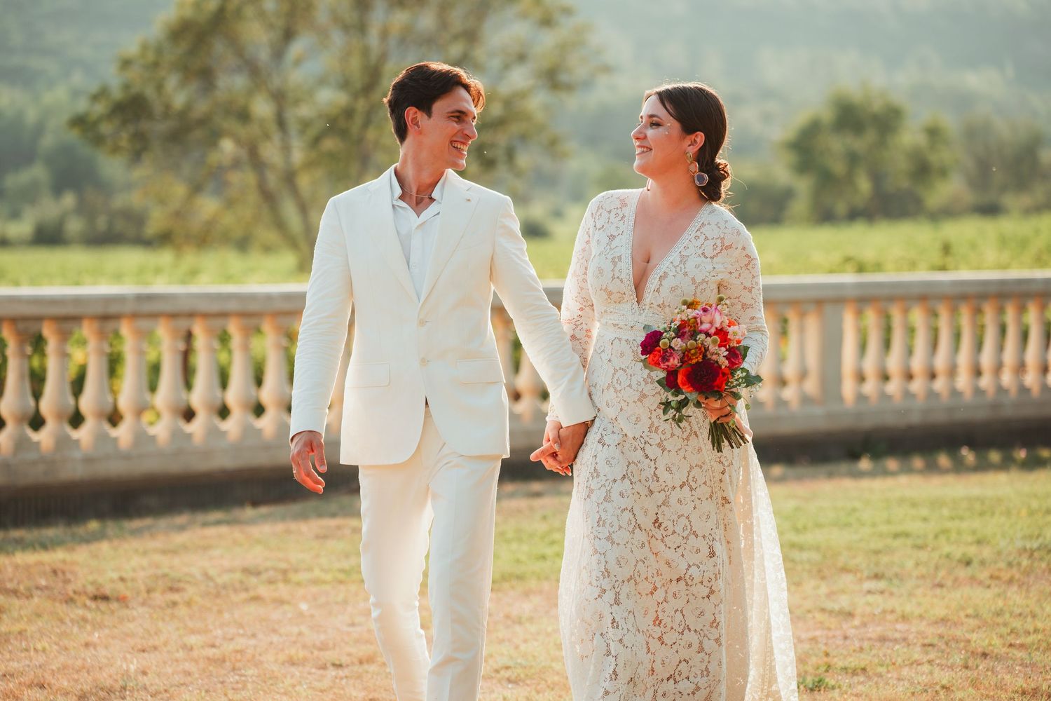 A couple in white wedding attire walks together through a sunlit garden terrace with scenic mountain views.