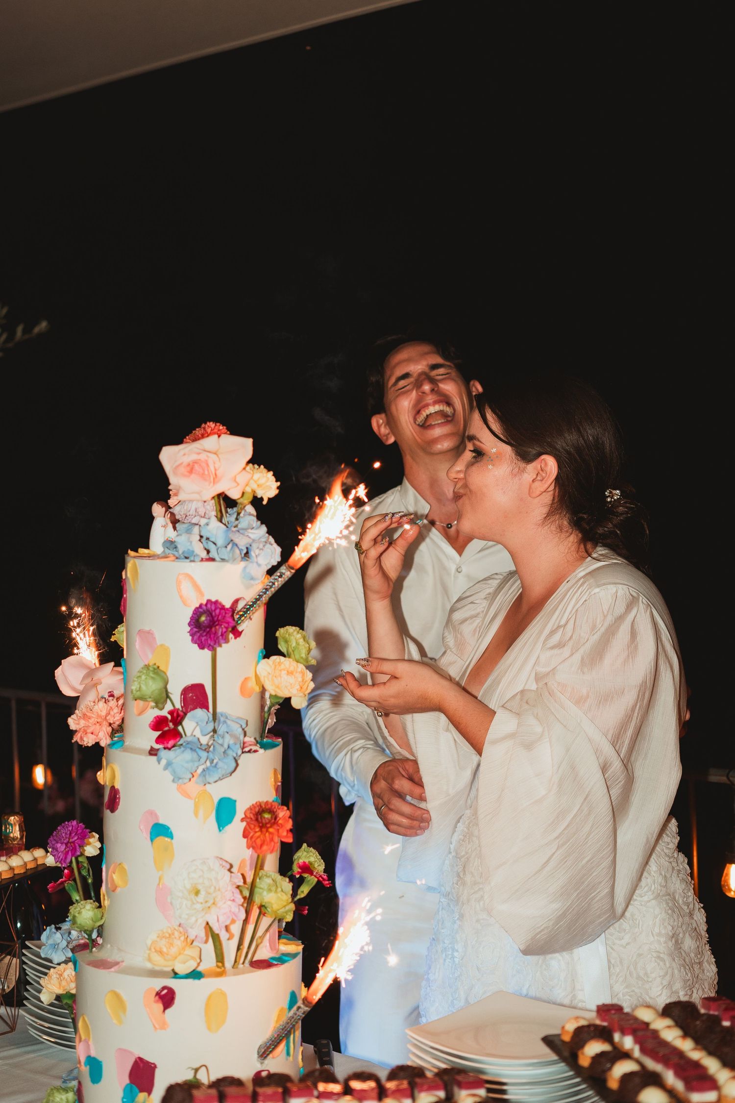 A colorful wedding cake decorated with bright flowers and butterflies is displayed next to a couple sharing a celebratory moment.