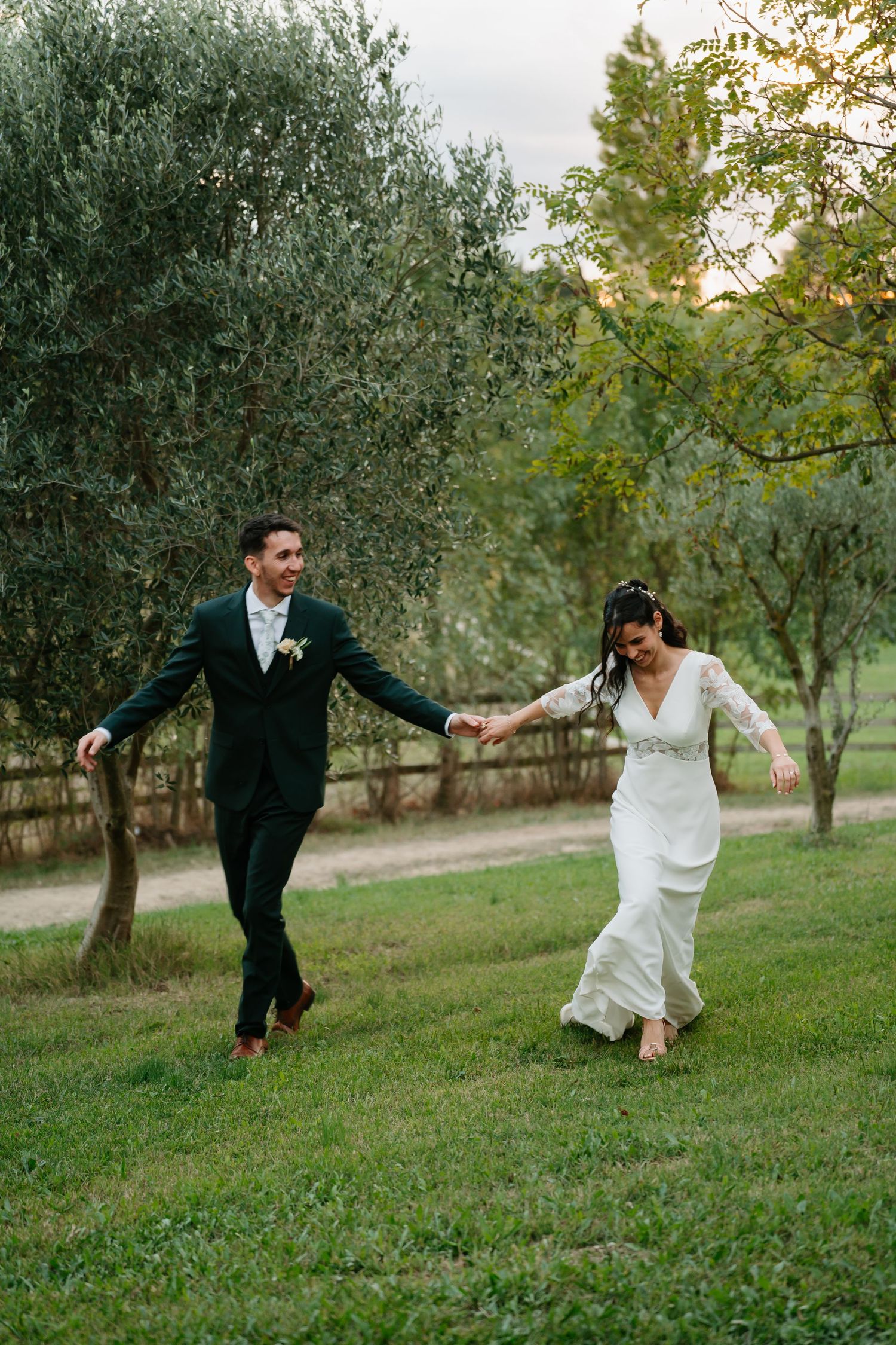 A couple playfully runs and holds hands in a scenic olive grove at sunset during their wedding celebration.