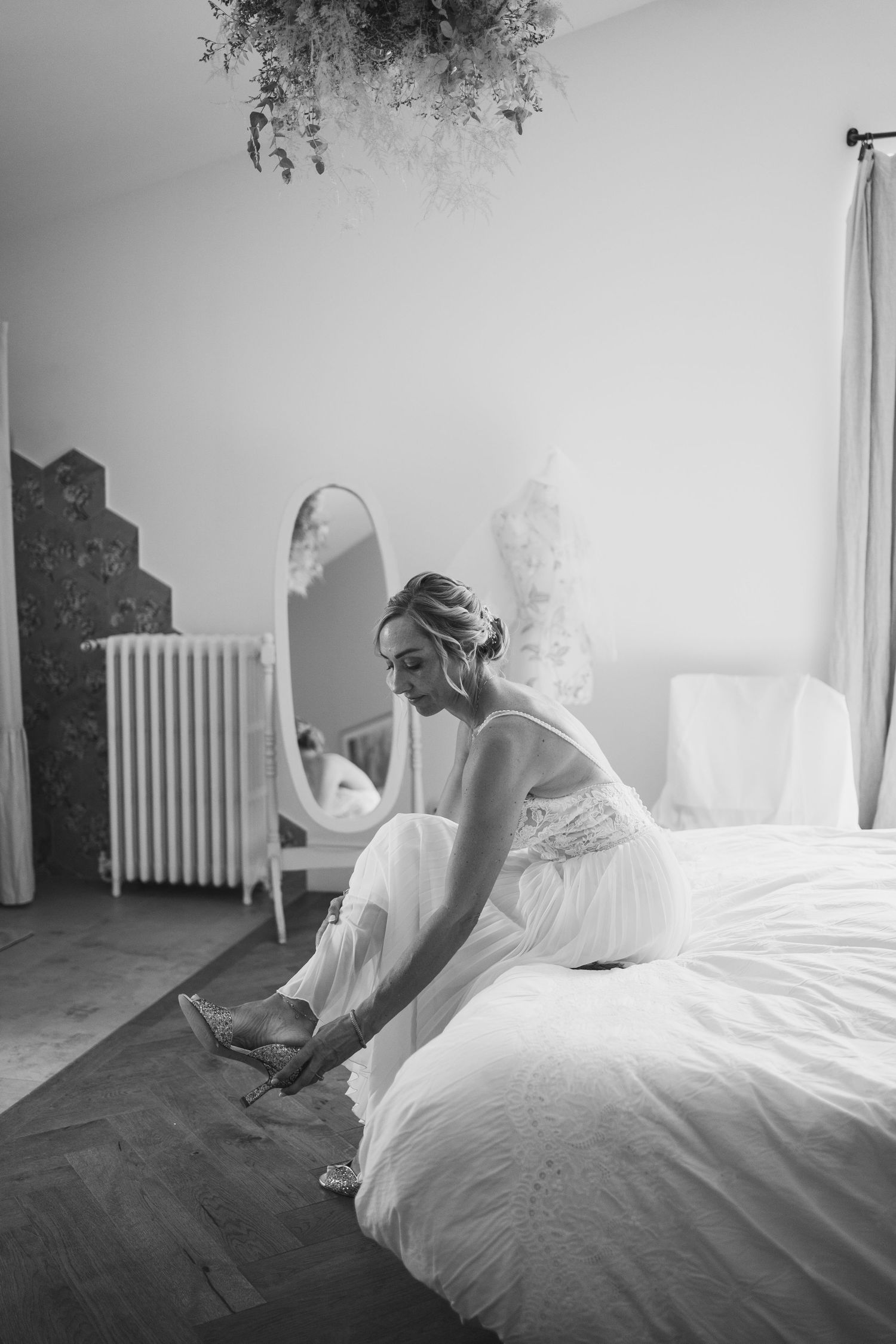 A bride in a white dress sits on a bed while putting on her shoes in a sunlit room with vintage decor.