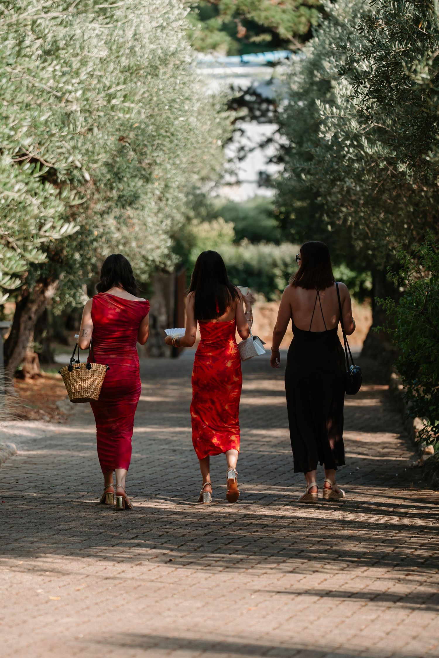 Three bridesmaids in red and black dresses walk down a tree-lined path during an outdoor wedding celebration.