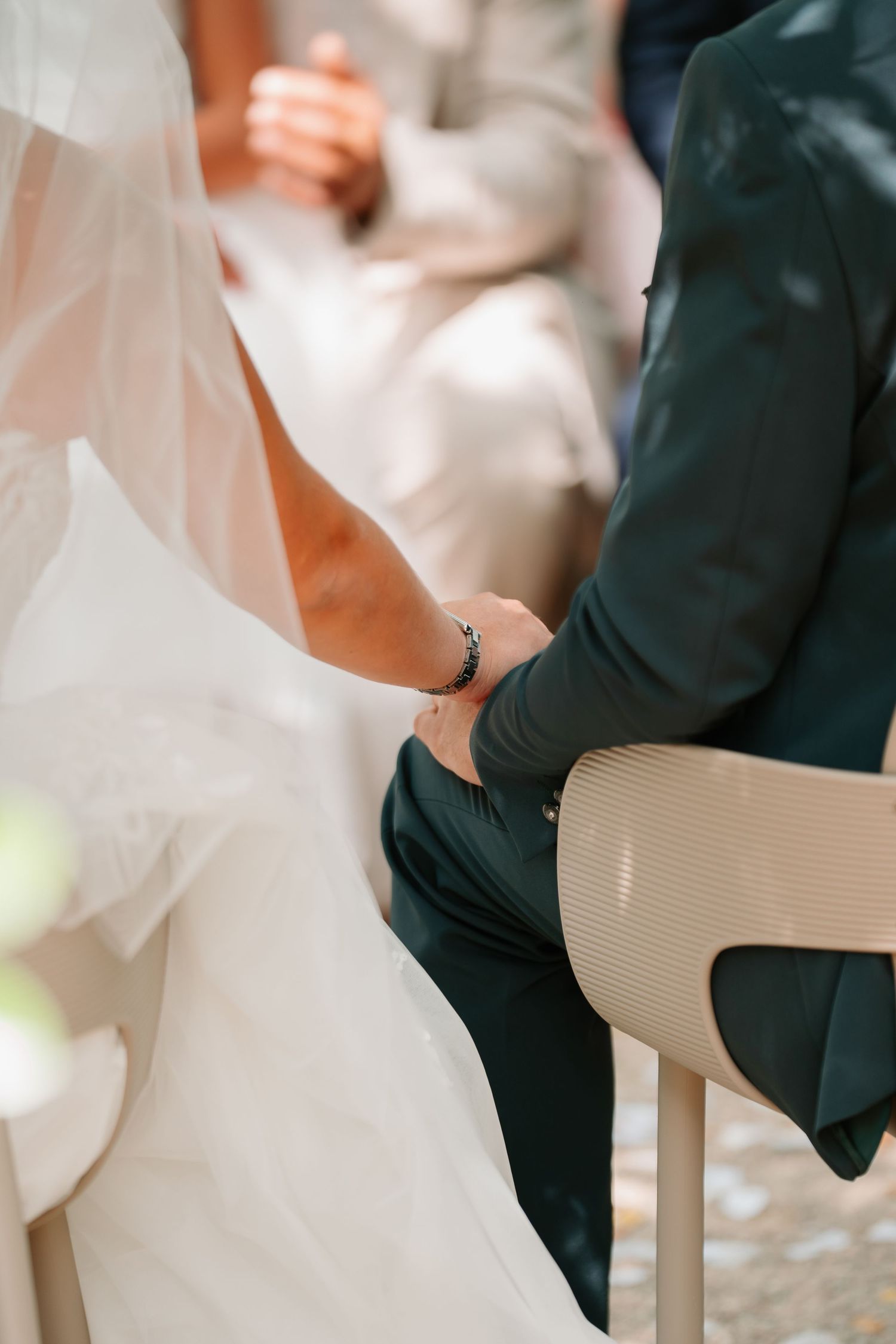 Close-up detail of bride in white dress and groom in dark suit sitting on white chairs during outdoor wedding ceremony.