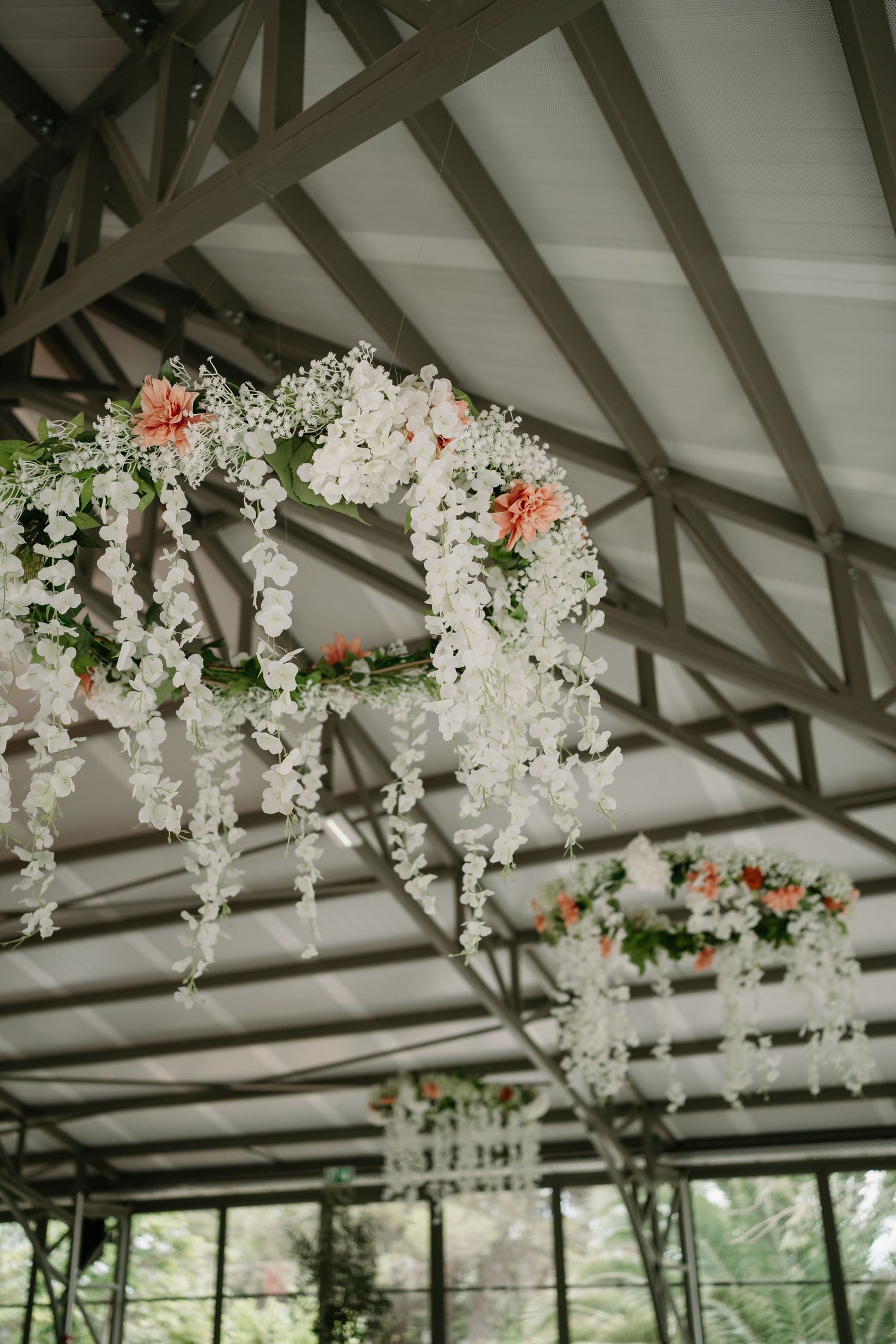Elegant white floral wedding arch with coral accents hangs from exposed wooden beams in an indoor venue.