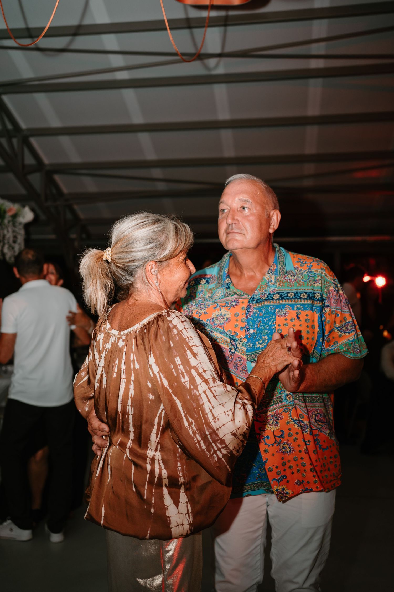 Couple dancing together at a dimly lit event with one person wearing a shiny brown jacket and another in a bright patterned shirt.