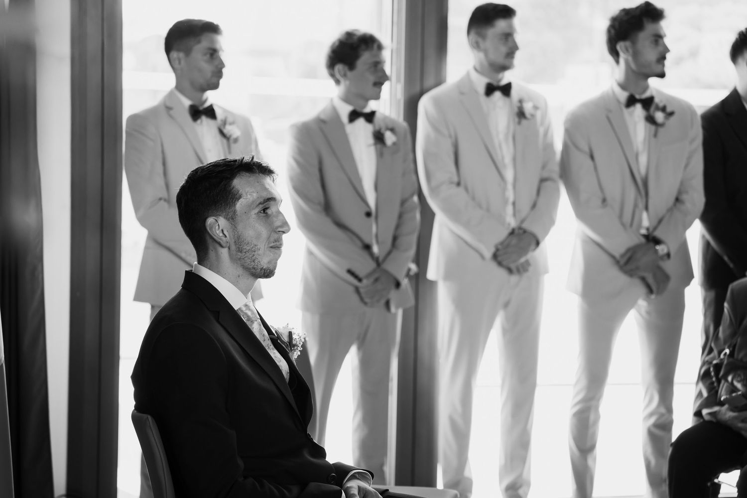 A black and white photo shows a group of men in formal attire standing in a row against a striped backdrop.