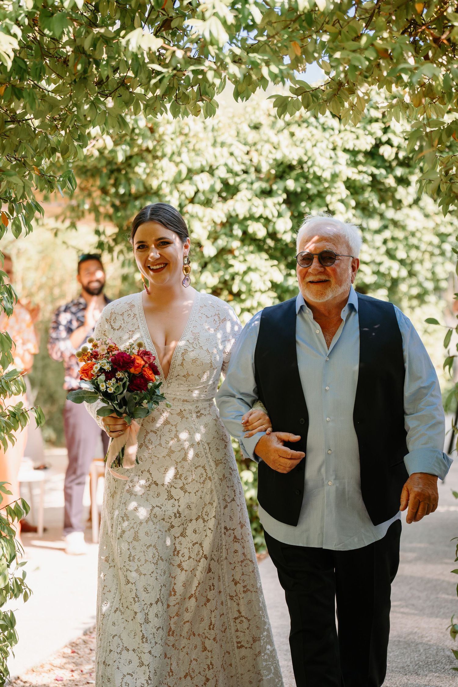 A bride in a white lace wedding dress walks down a garden path holding a colorful bouquet of flowers.