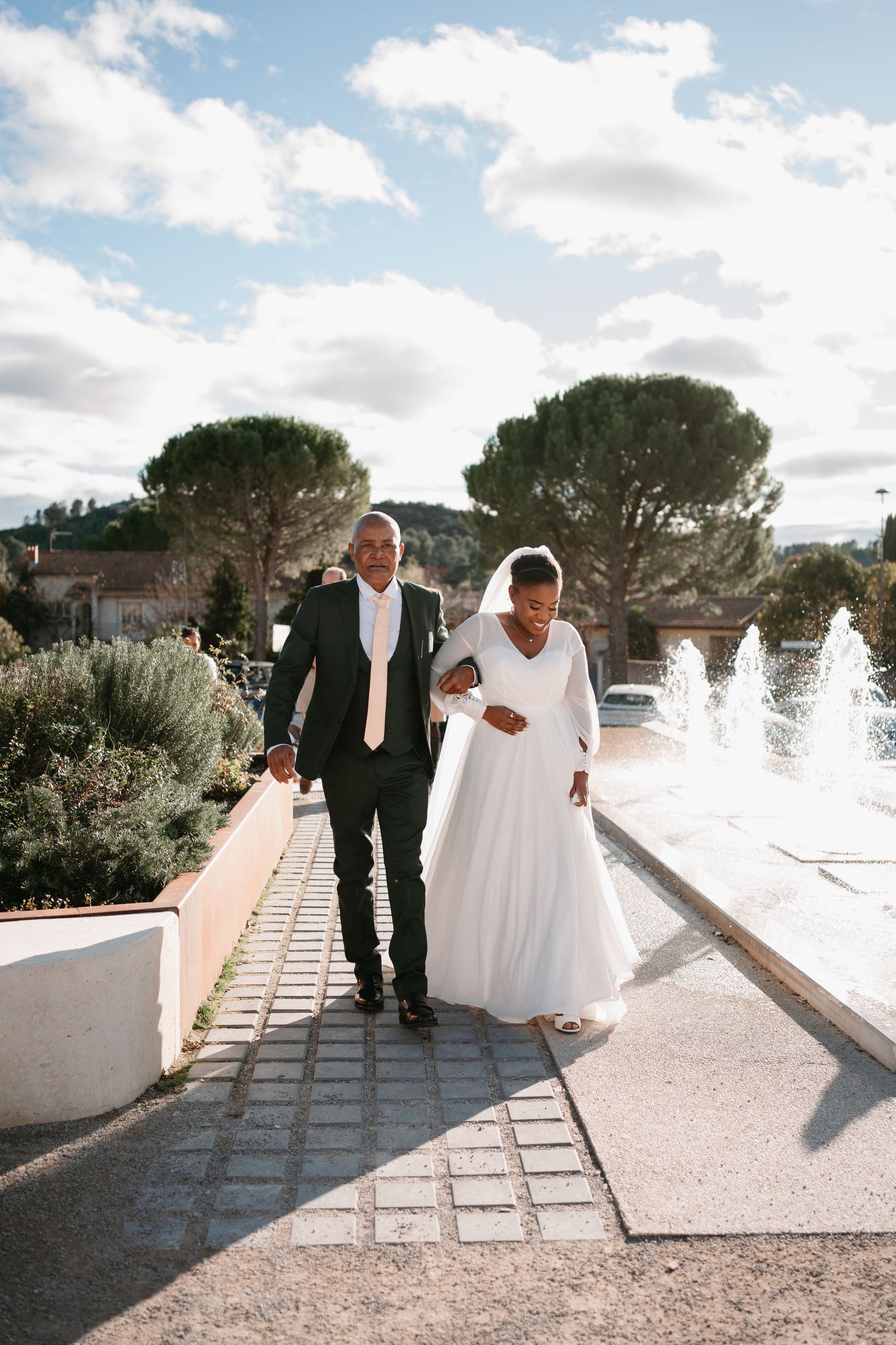 Wedding party walks along white Mediterranean villa pathway under bright blue sky.