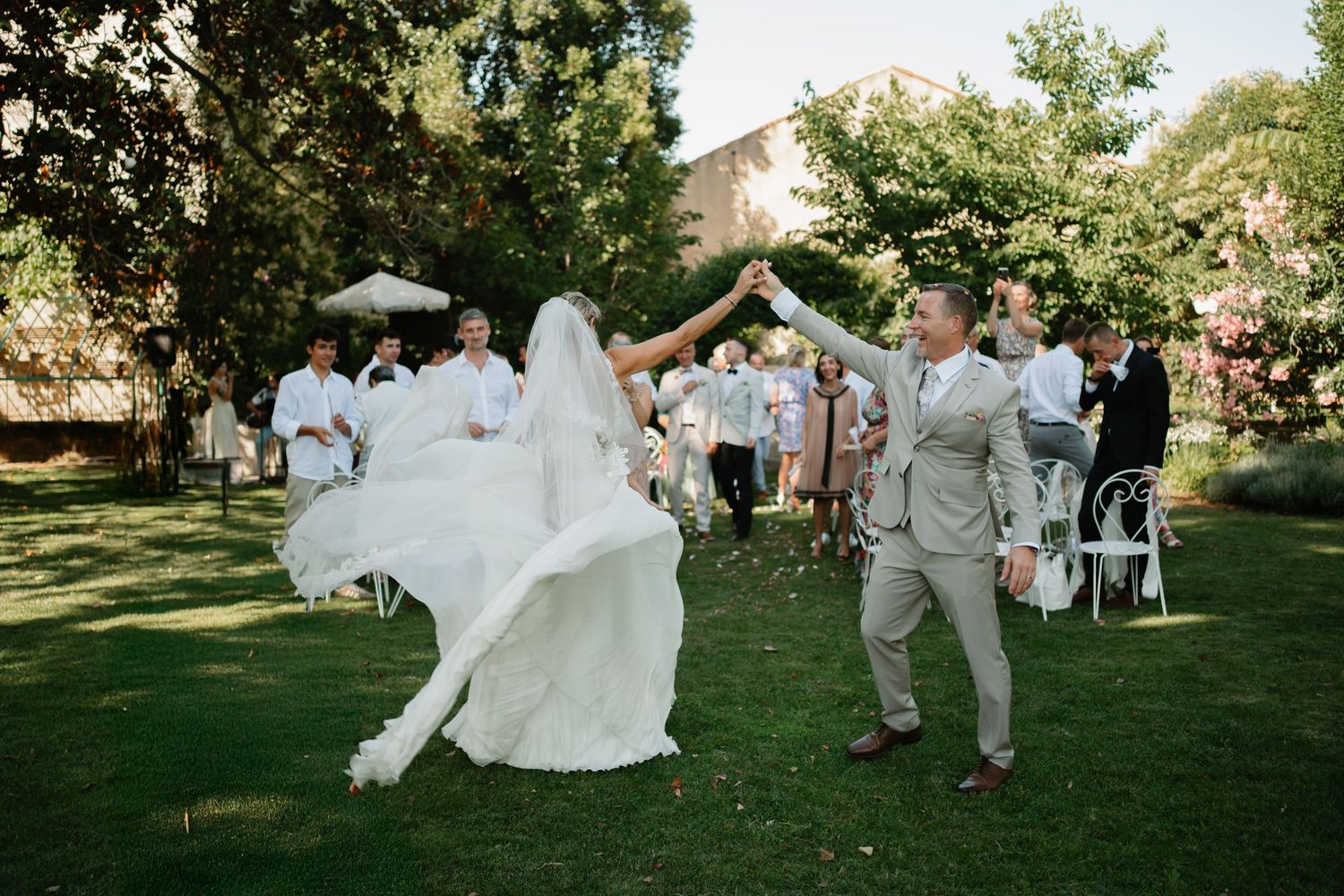 Wedding party members dance and celebrate together on a lush green lawn during an outdoor summer reception.