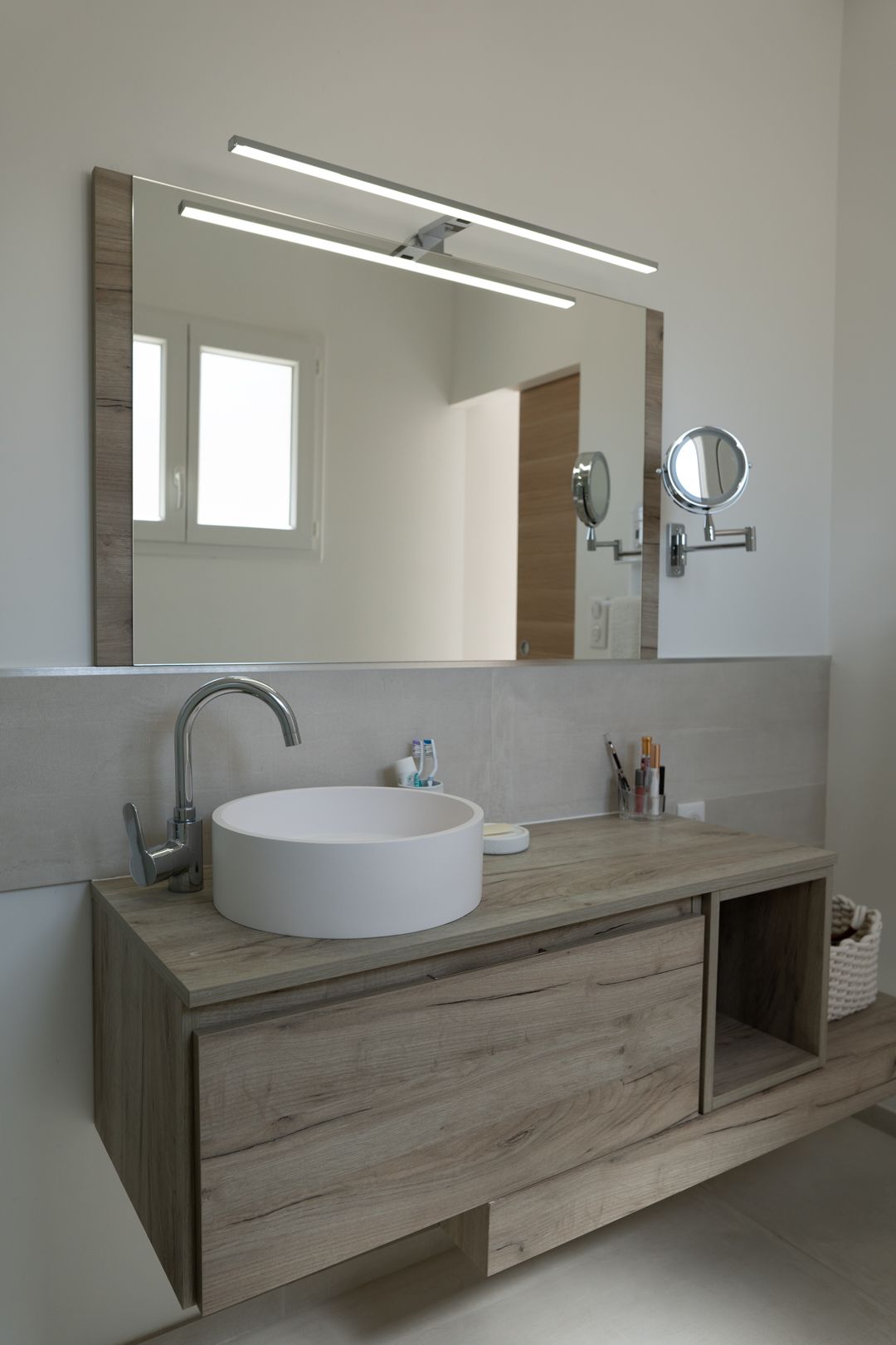 Modern bathroom interior with floating wooden vanity, white vessel sink, and large mirror illuminated by LED lighting.