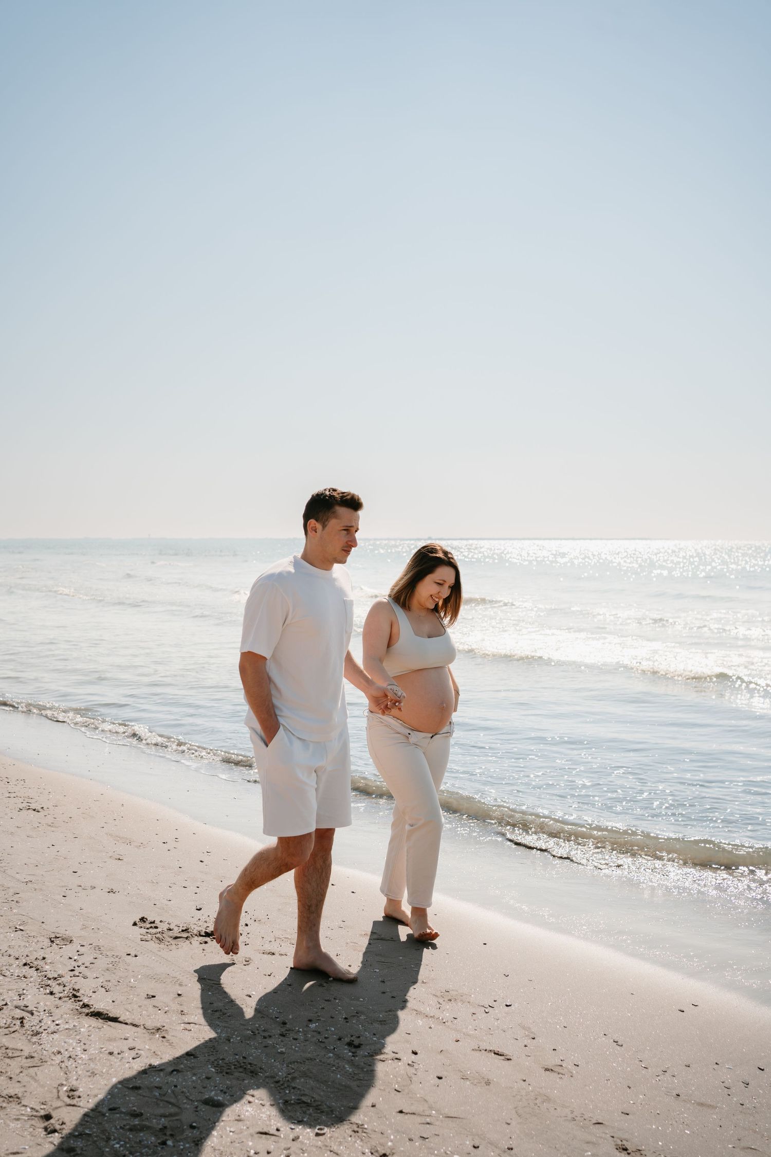 Couple walking barefoot on sunny beach while expecting mother touches her pregnant belly.