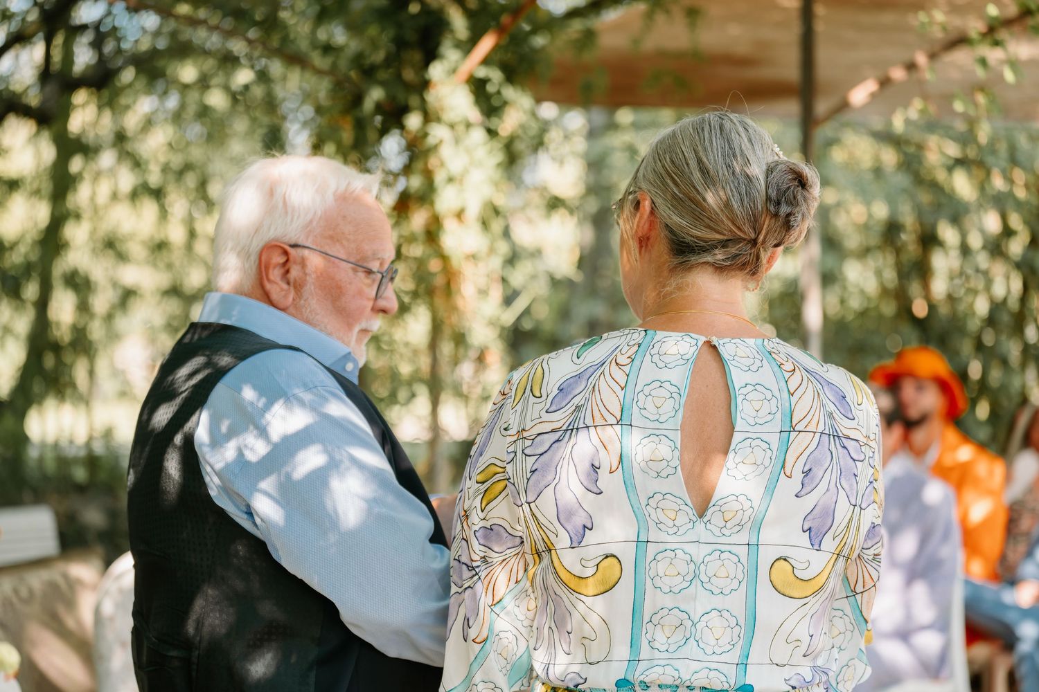 A group of people enjoy a sunny outdoor gathering with white flowers and decorative elements in the background.