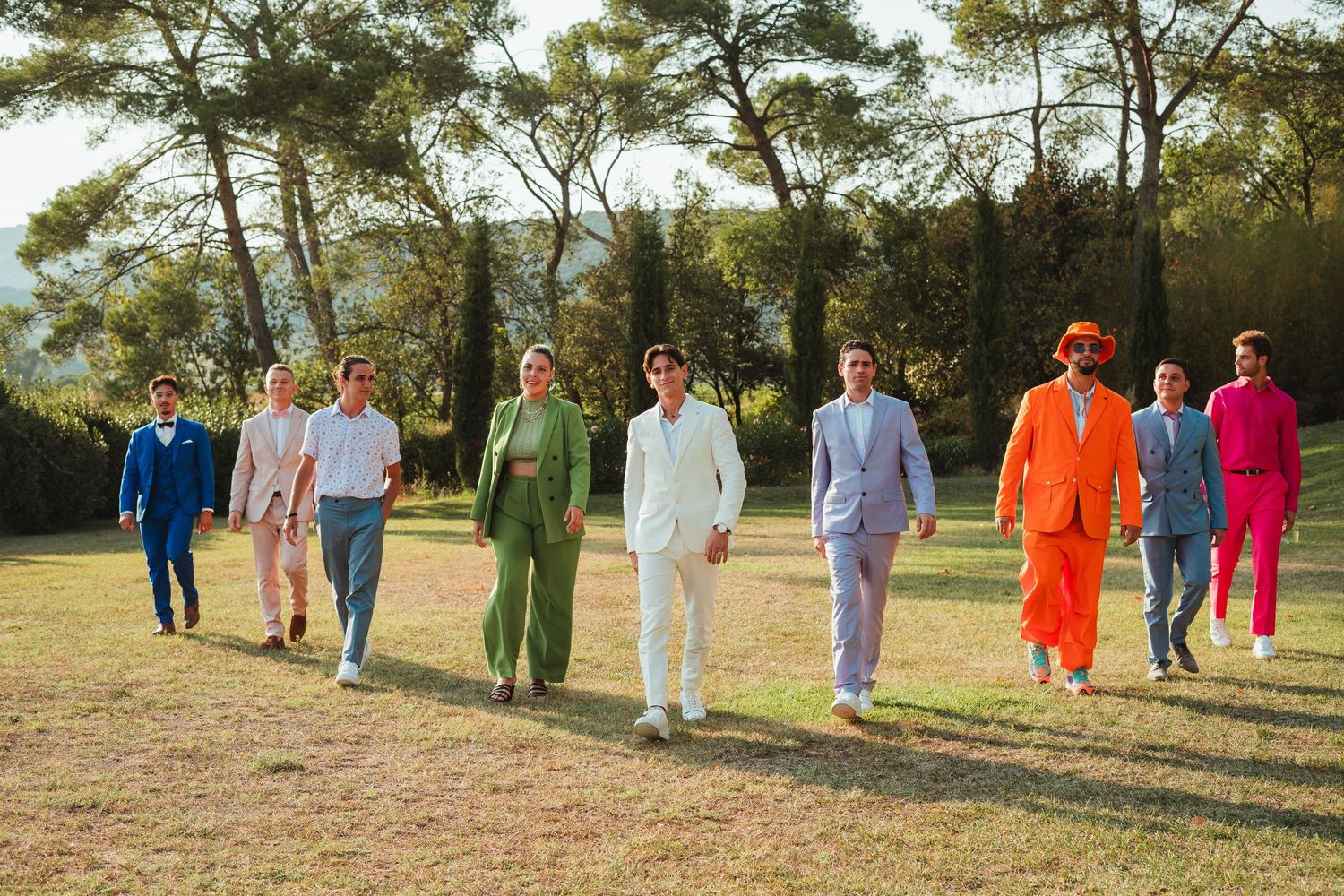 A group of people wearing colorful suits walk in a line on a grassy hill with pine trees in the background.