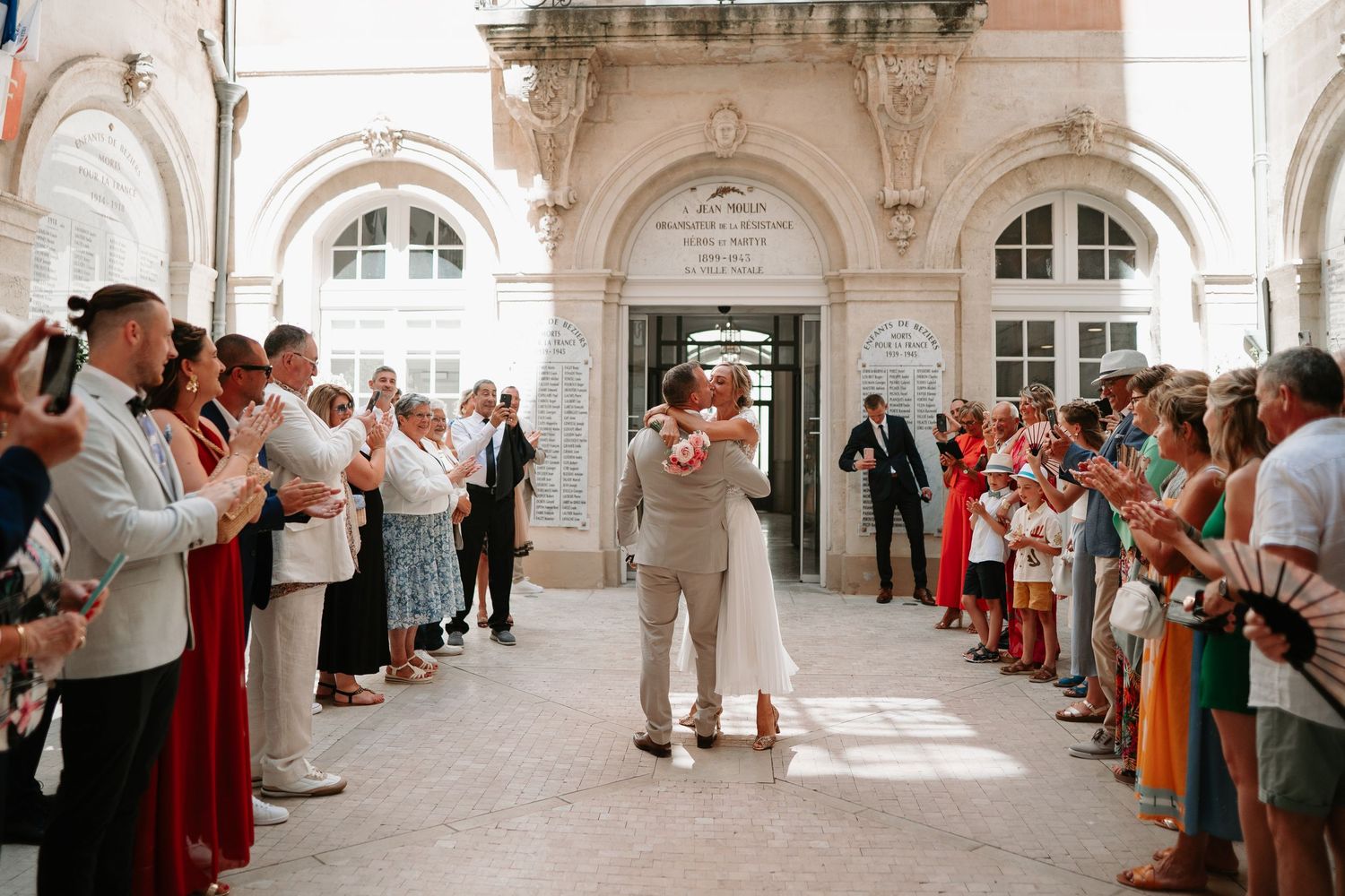 A group of people in traditional attire stand in a line against ornate white stone archways during a ceremony.