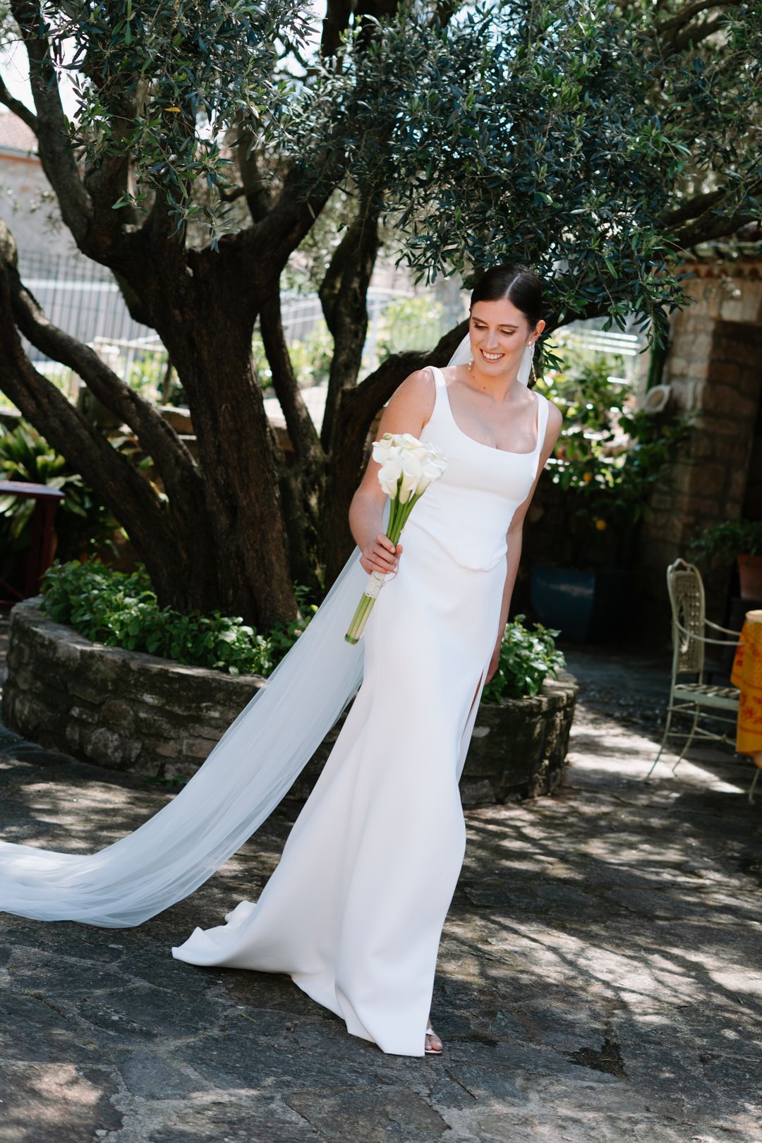 Bride in elegant white gown holding calla lily bouquet with flowing veil, standing beneath olive tree in garden.