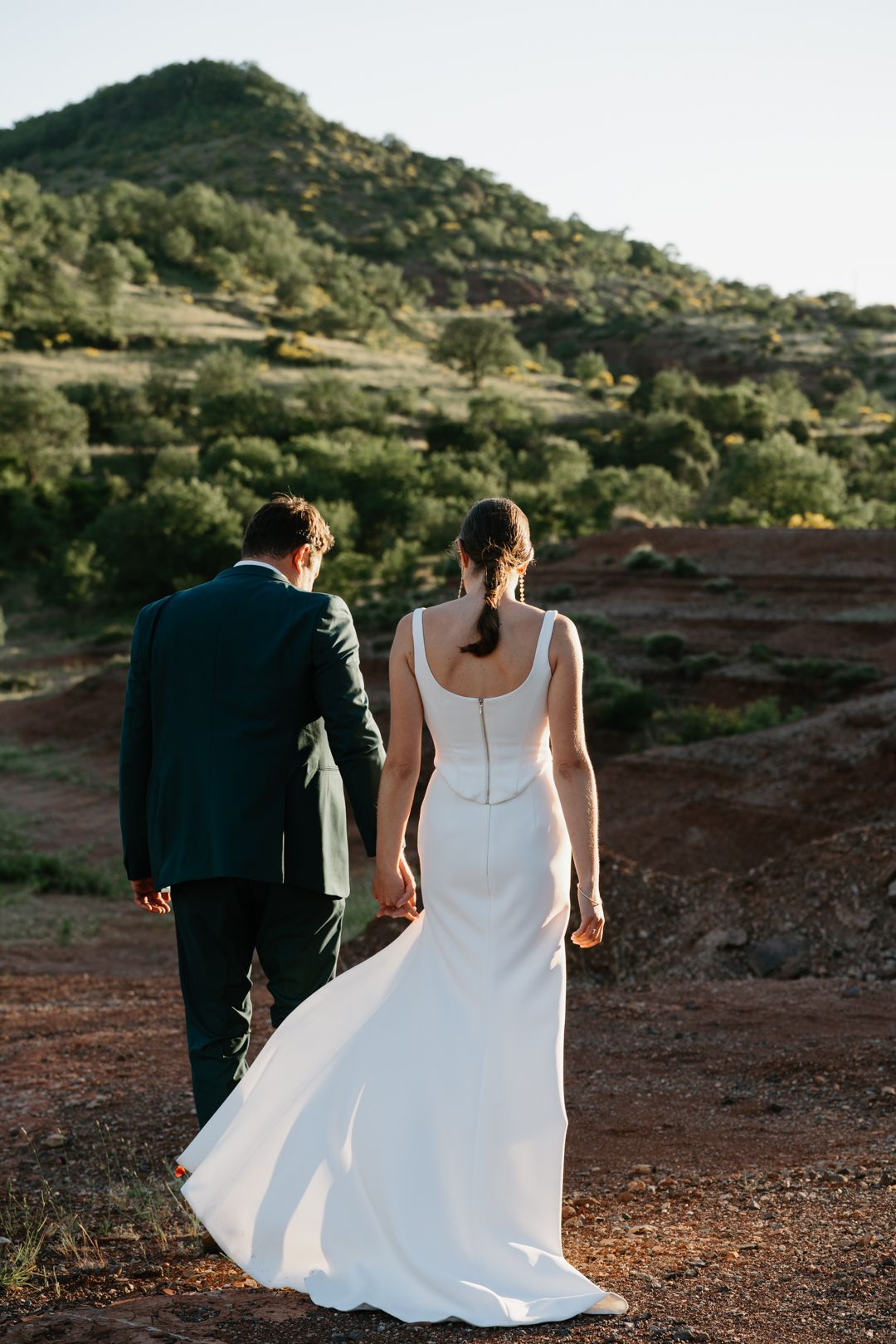 Bride and groom holding hands walking toward lush green hills at golden hour in a desert landscape.