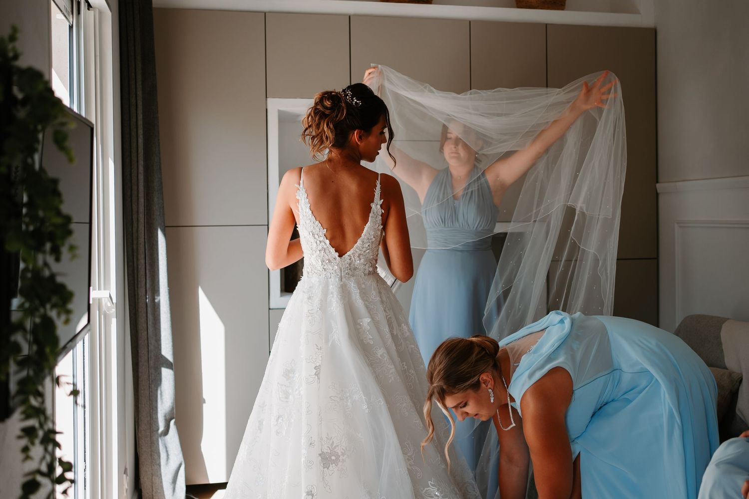 Wedding dress preparation scene in sunlit room with tulle veil.