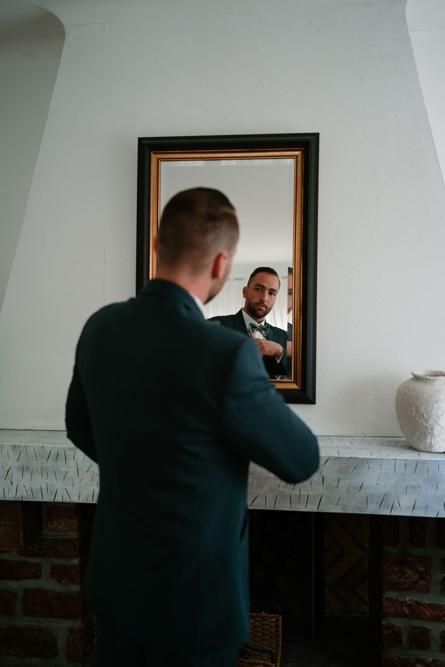 Two people in dark suits interact with a framed mirror mounted on a white wall with exposed brick detail.