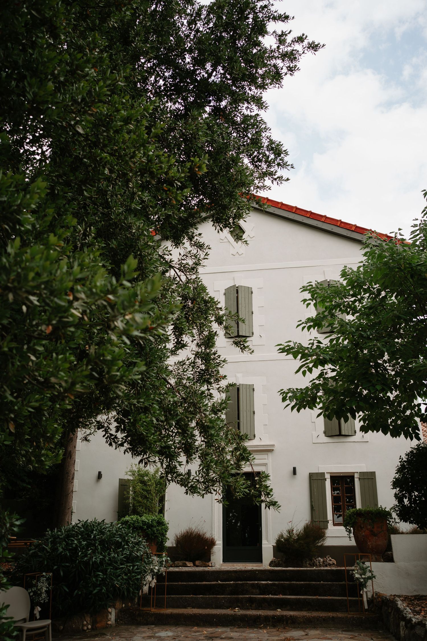 White Spanish colonial style house with red tile roof and green trees surrounding the entrance walkway.