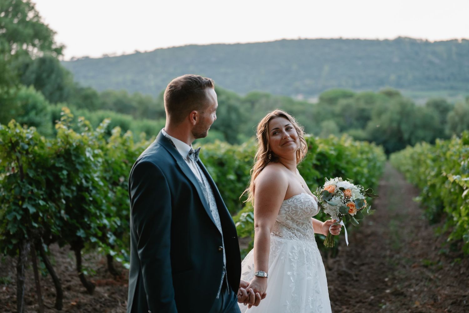 A couple stands together in a lush vineyard with rolling hills and greenery in the background during sunset.