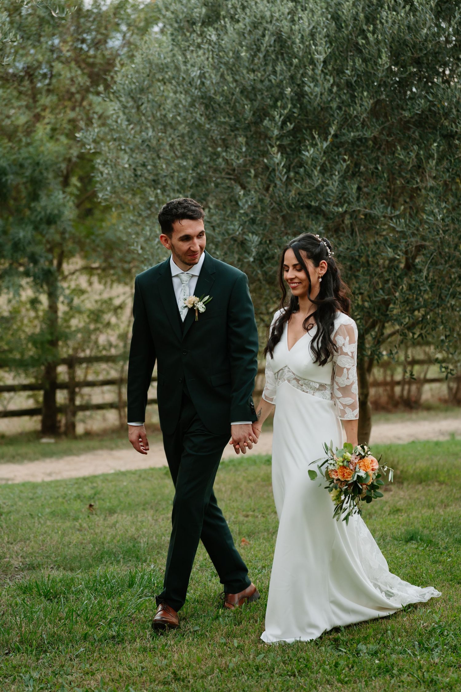 A couple walks hand in hand through an olive grove on their wedding day, with the bride holding a vibrant autumn bouquet.