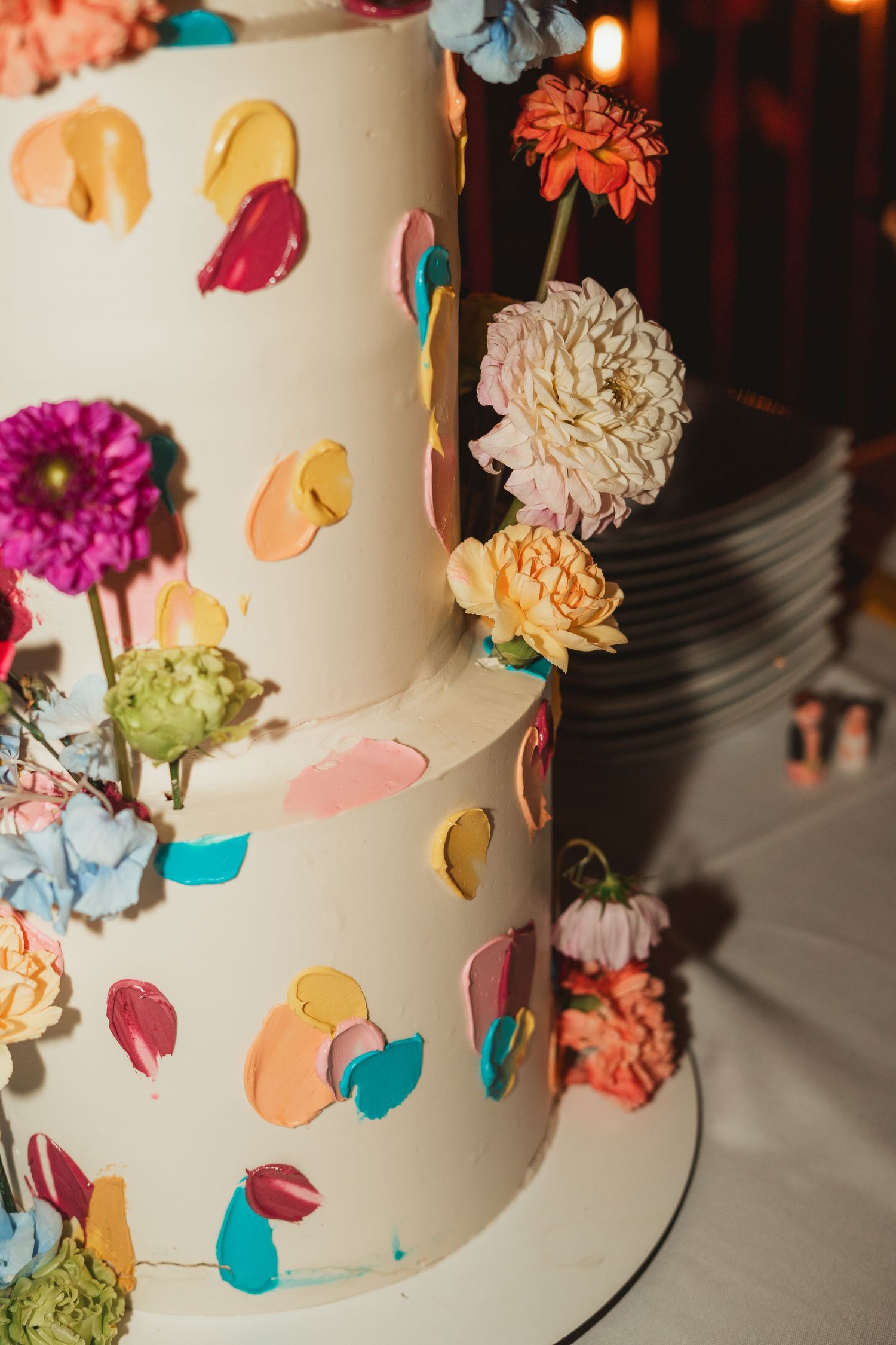 Close-up view of a white wedding cake decorated with colorful fresh flowers and petals in turquoise, pink and orange tones.