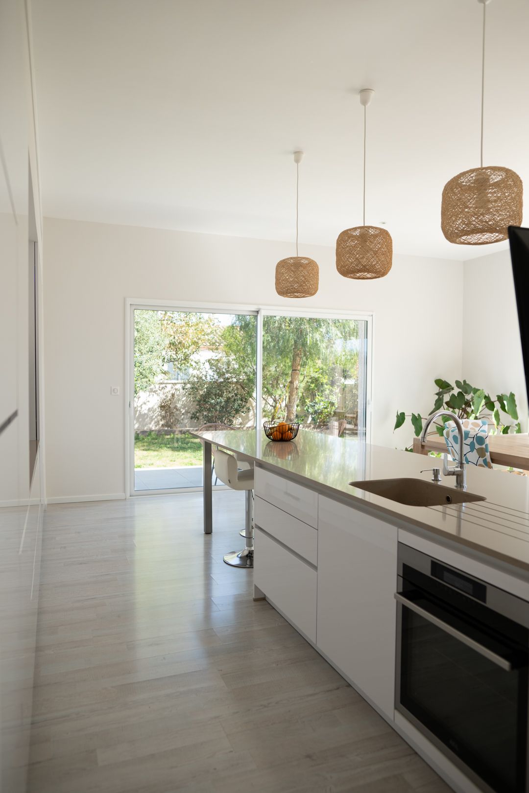 Modern white kitchen with wicker pendant lights, sleek cabinets, and large windows offering natural light and garden views.