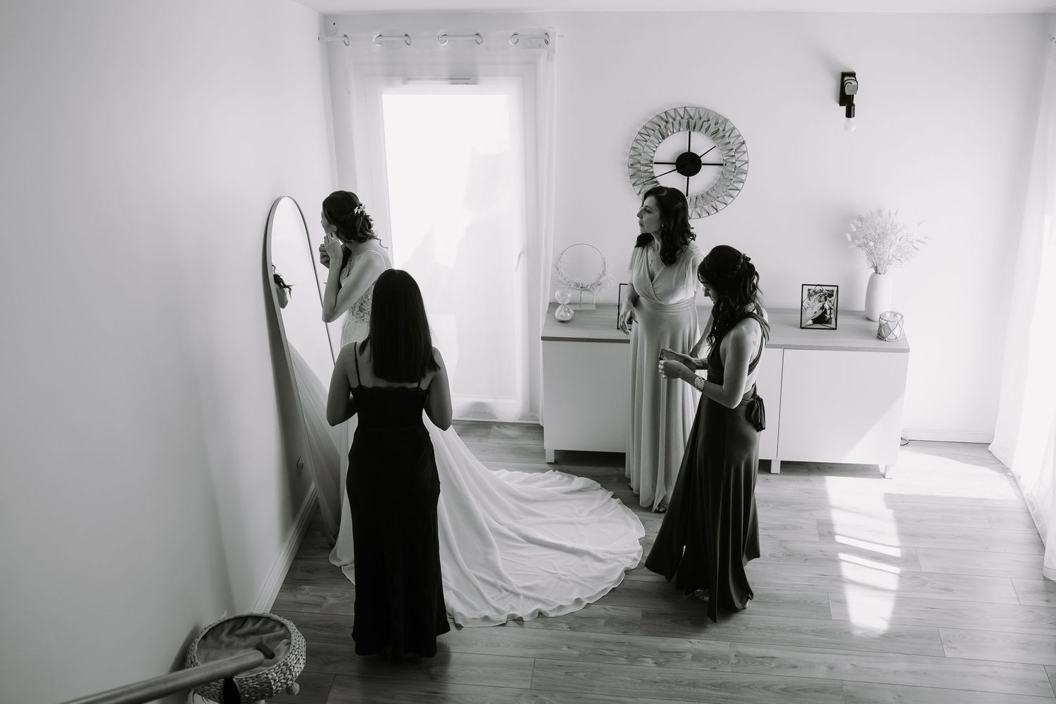 Black and white photo of a bride in a white gown with bridesmaids helping her prepare in a bright, elegant room.