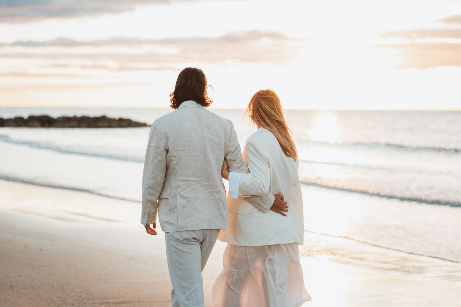 A couple walking along a beach at sunset wearing light colored clothing.