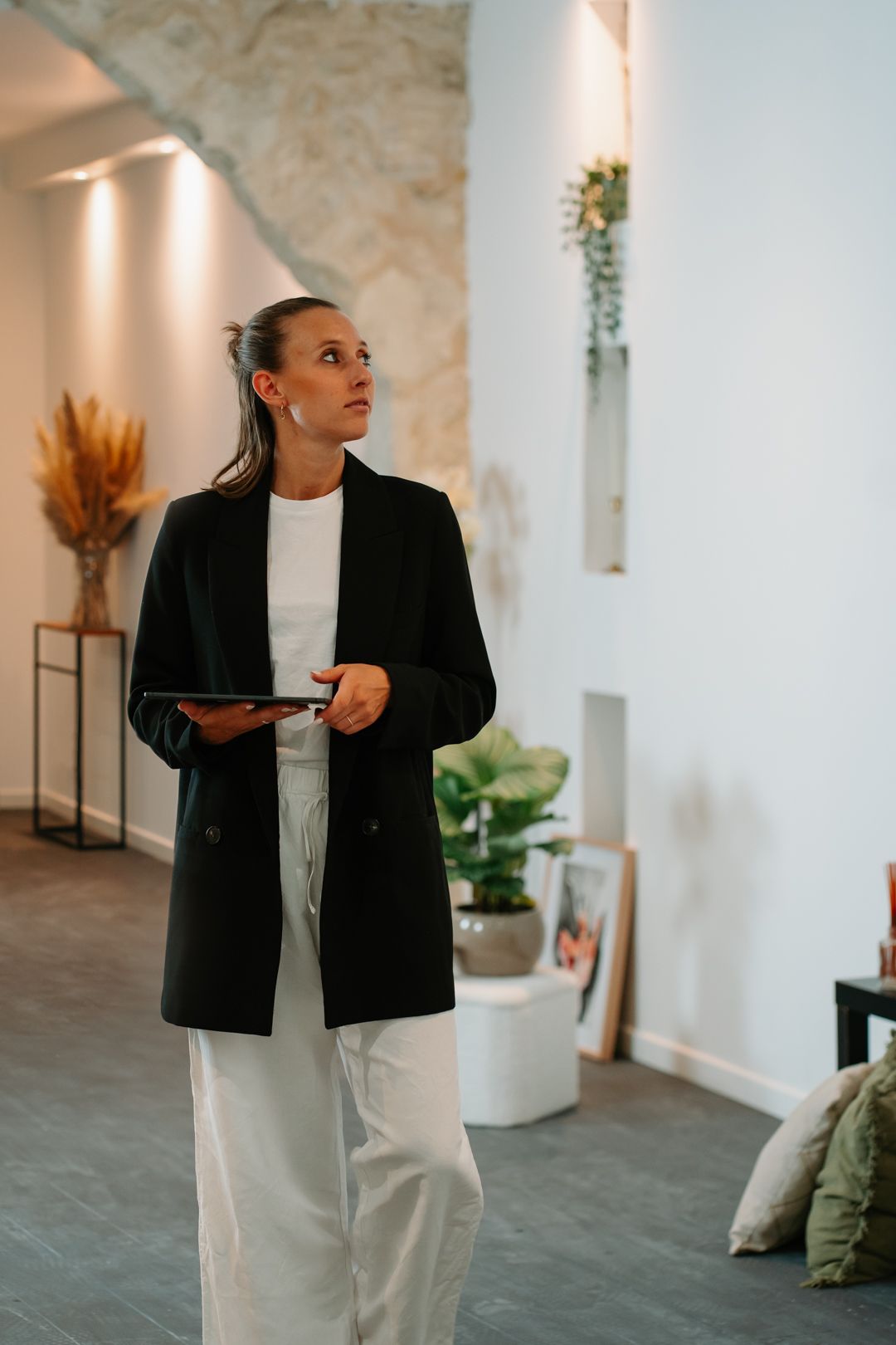 Professional wearing a black blazer and white pants stands in a modern minimalist space with natural decor elements.