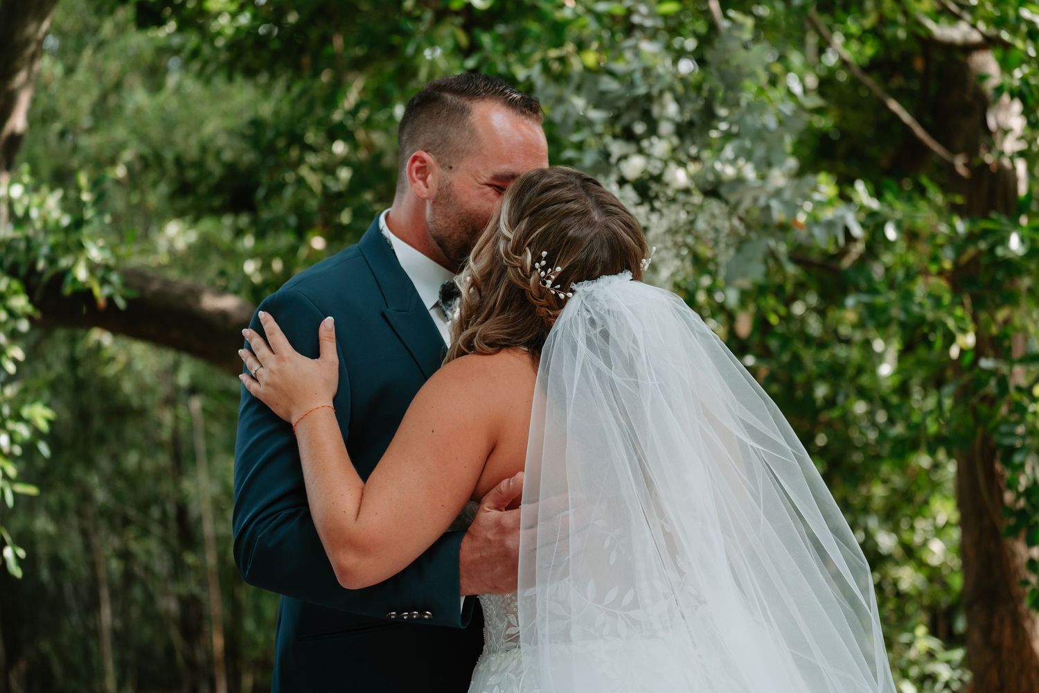 A tender moment between newlyweds embracing outdoors with a flowing white veil against a green garden backdrop.