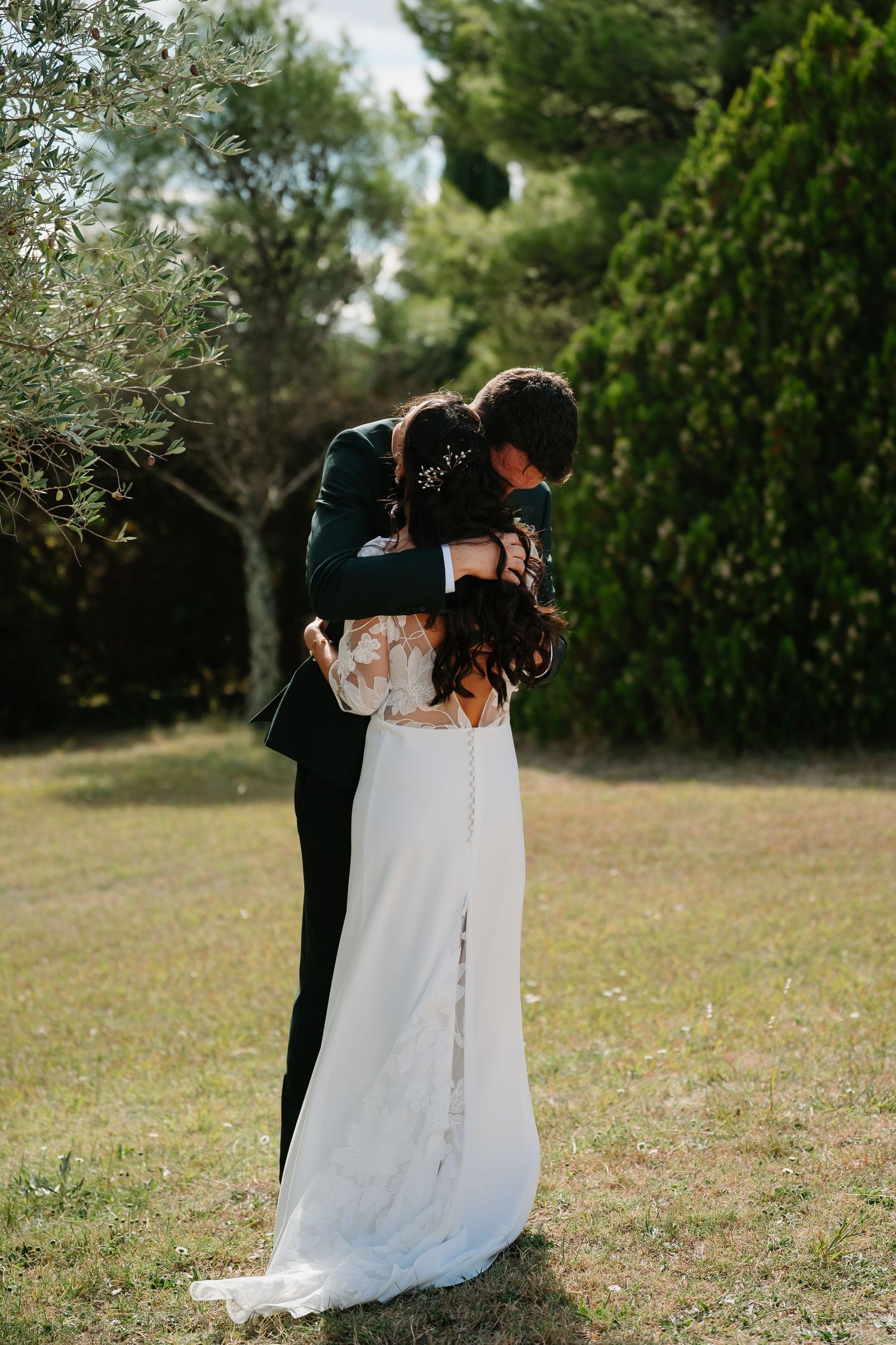 A romantic wedding couple embraces outdoors in a natural garden setting with green trees in the background.