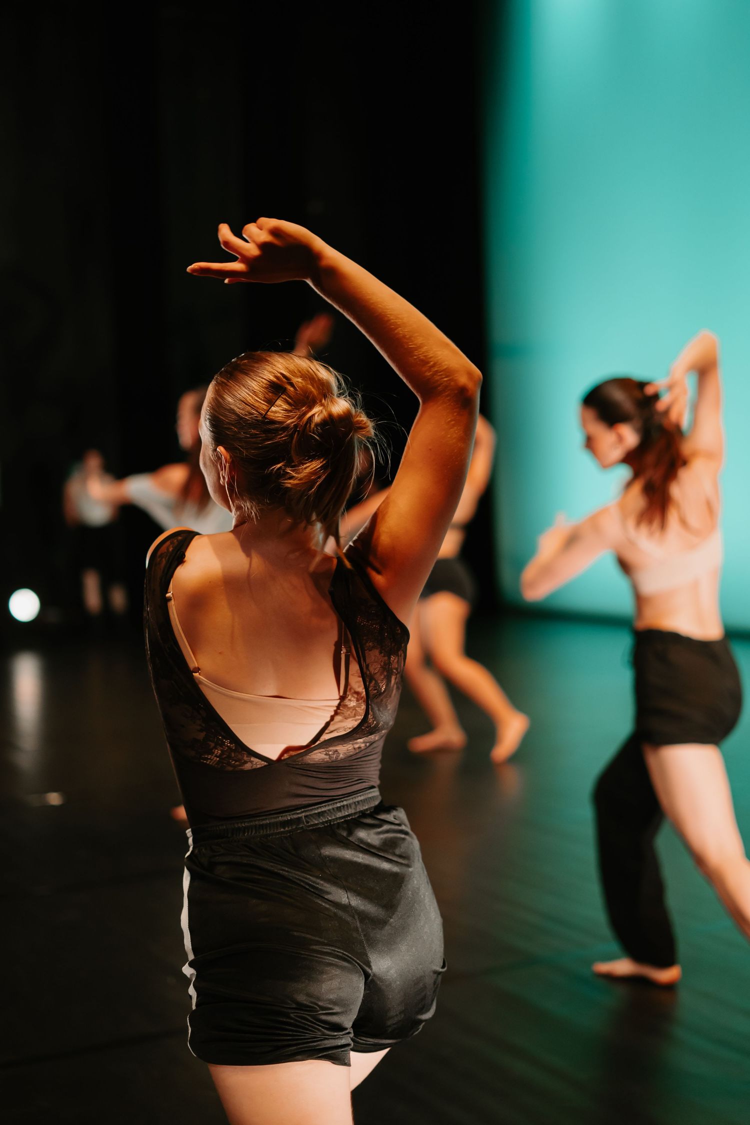 Modern dancers perform in black attire against a turquoise backdrop on a dark stage during a contemporary dance show.
