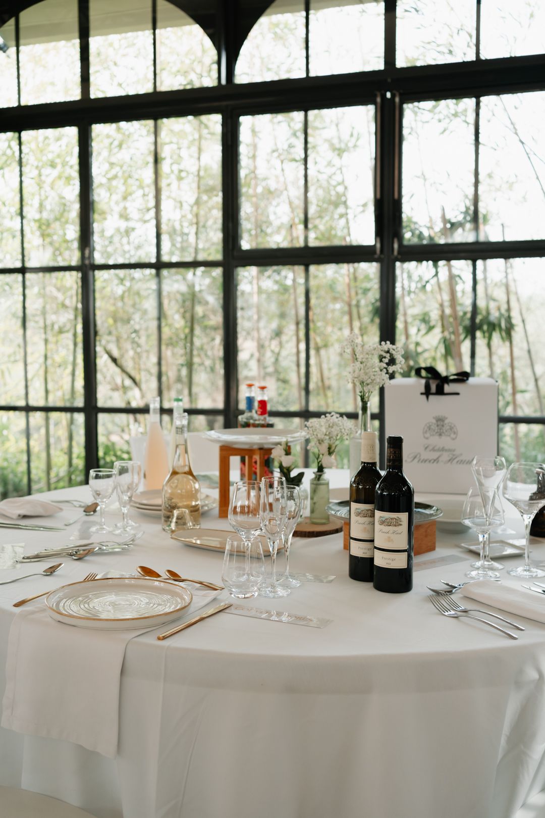 Formal dining tables set with white linens, wine glasses and bottles in a bright room with large windows.