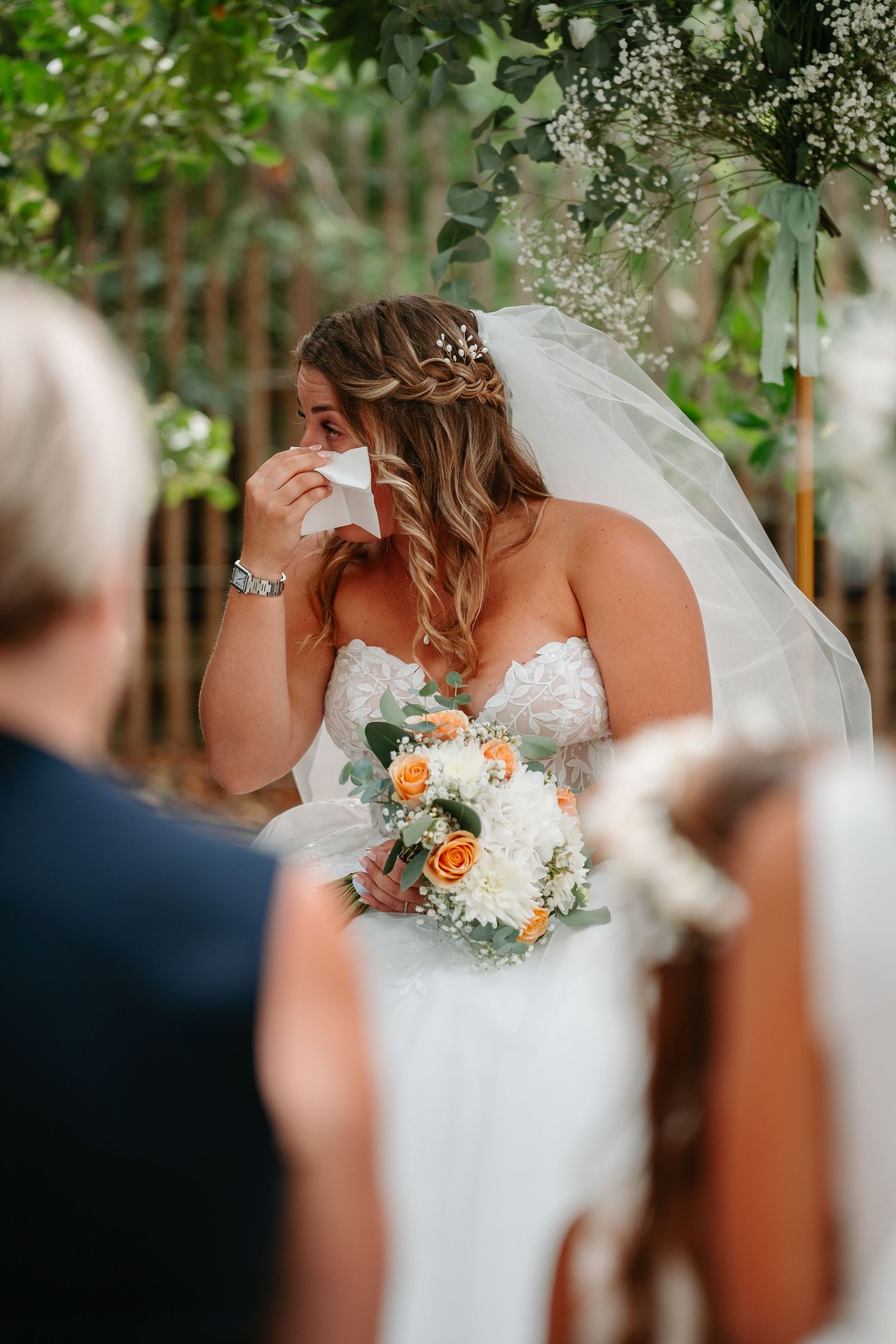 Bride in a white lace wedding dress holds a beautiful bouquet of orange roses and white flowers during an outdoor ceremony.