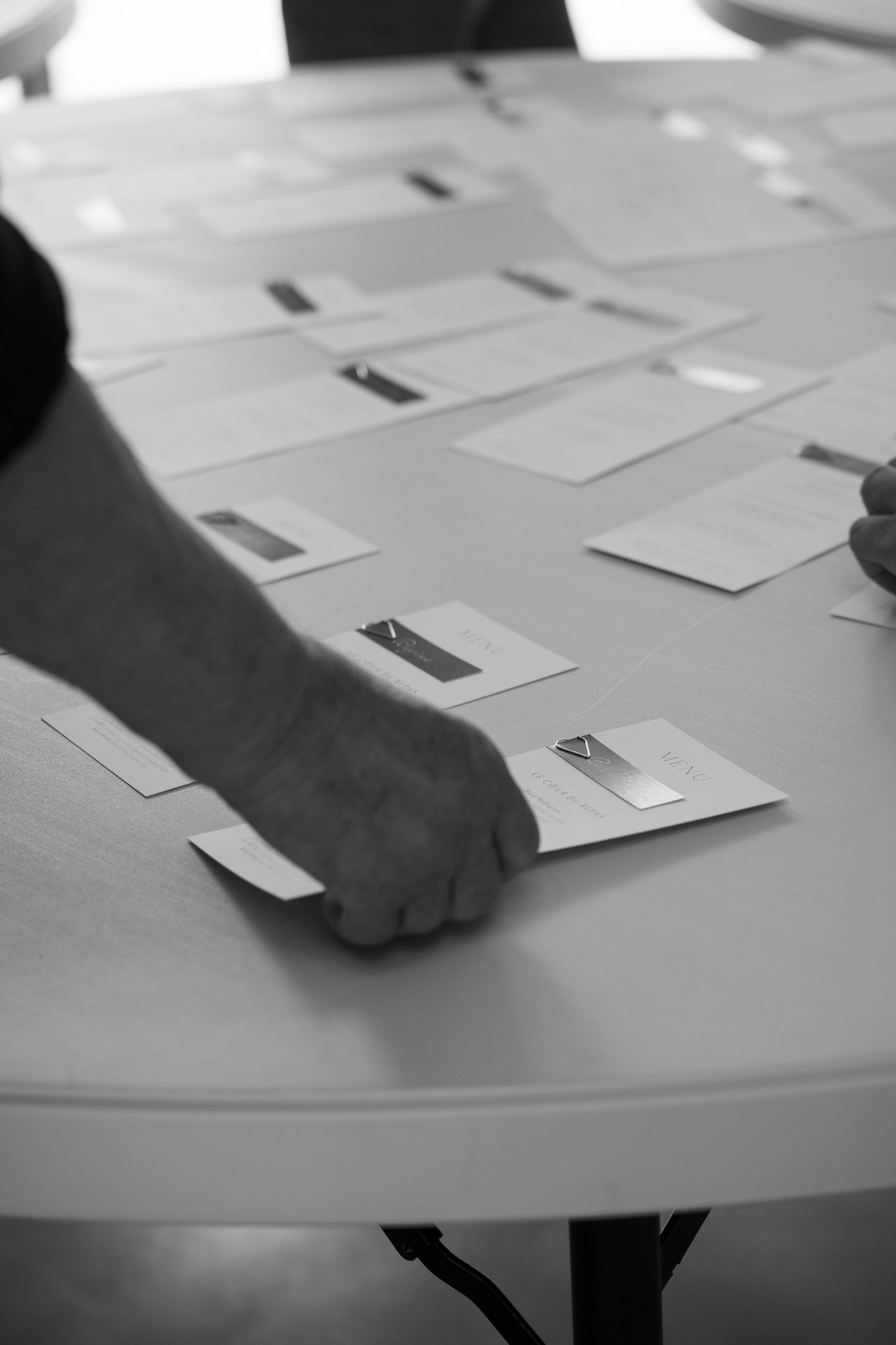 Black and white image showing hands arranging white cards or papers on a round table surface.