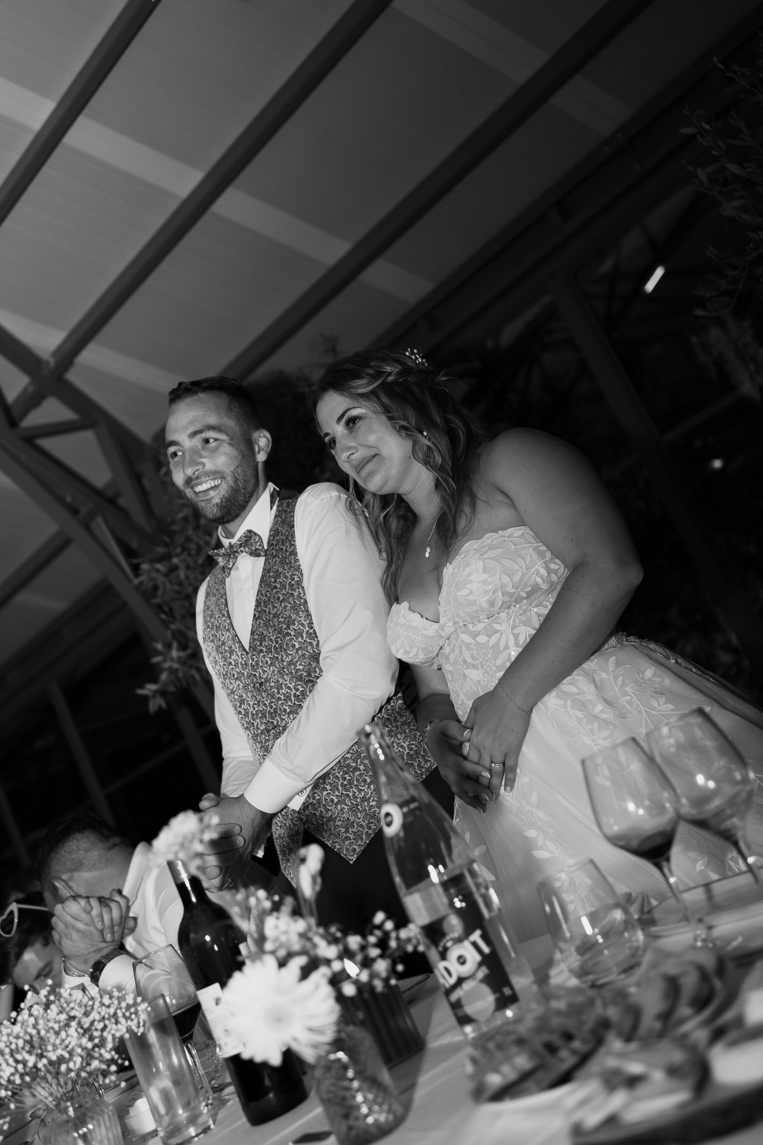 A black and white wedding reception photo showing a couple seated at a decorated table during their celebration.