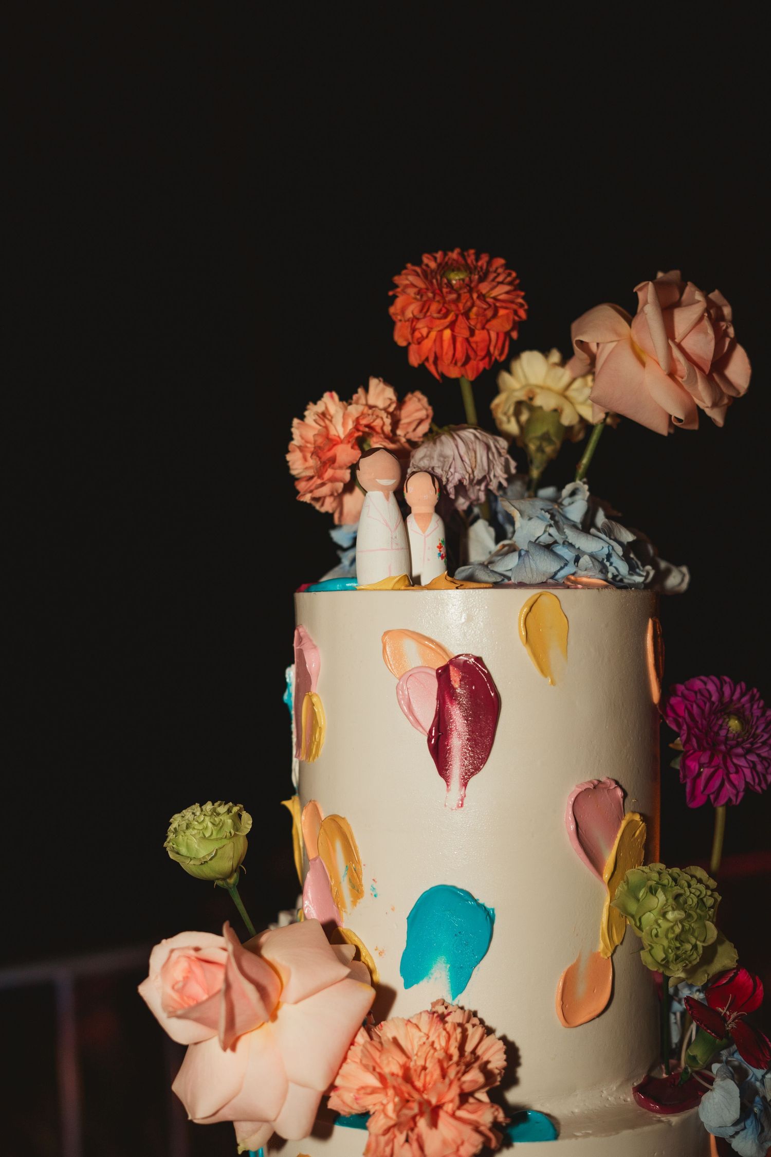 Elegant white wedding cake decorated with colorful dripping details and fresh flowers against a dark background.
