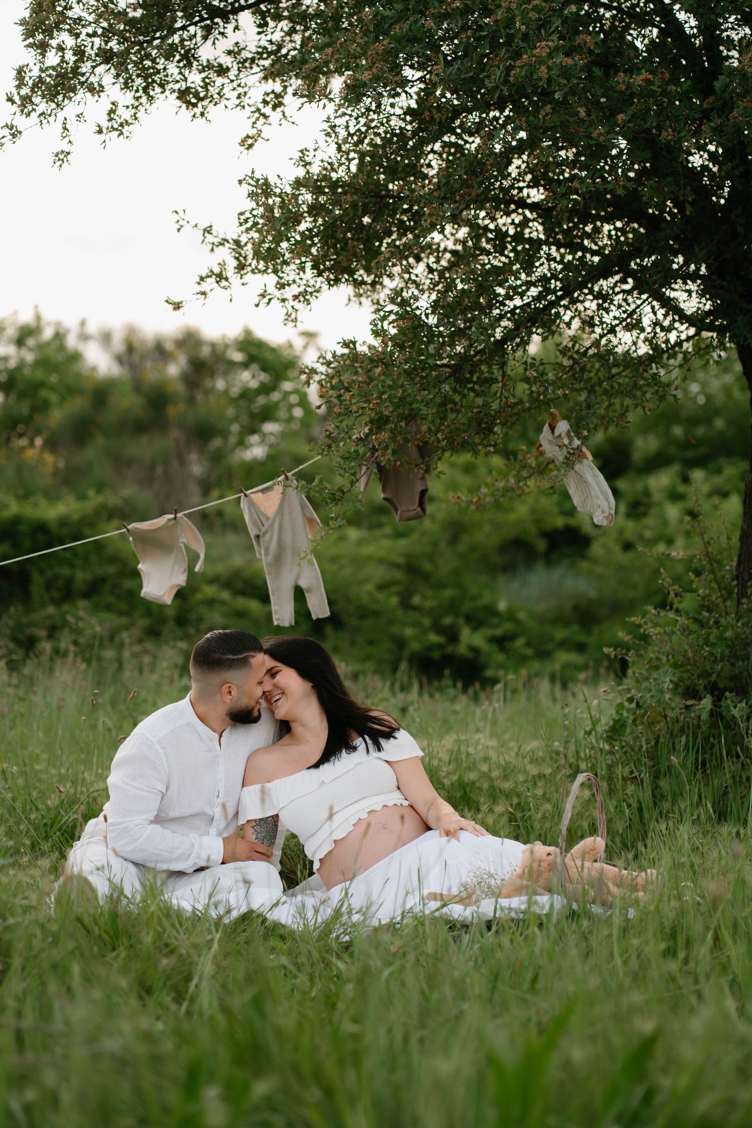 Couple in white clothing sits on grass with baby clothes hanging on clothesline above during outdoor maternity photoshoot.