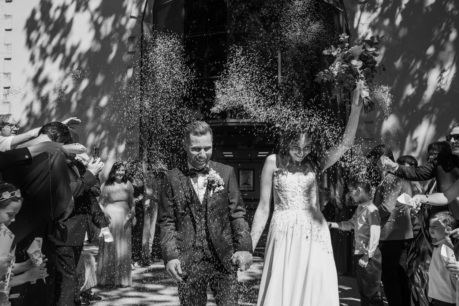 Black and white photo of a joyful newlywed couple exiting a church as guests throw confetti.