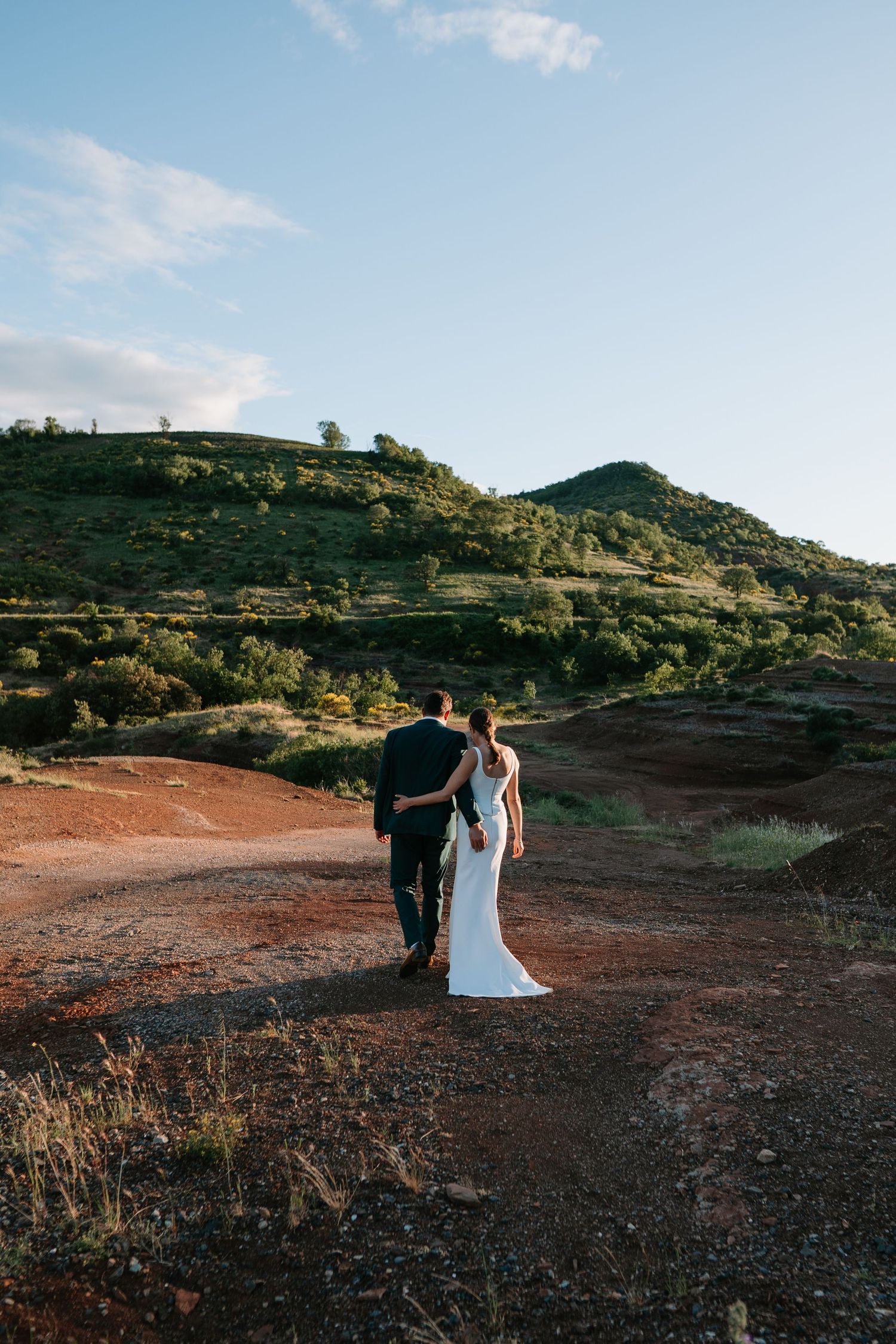 Bride and groom walk hand in hand on red dirt path with lush green hills under blue sky.