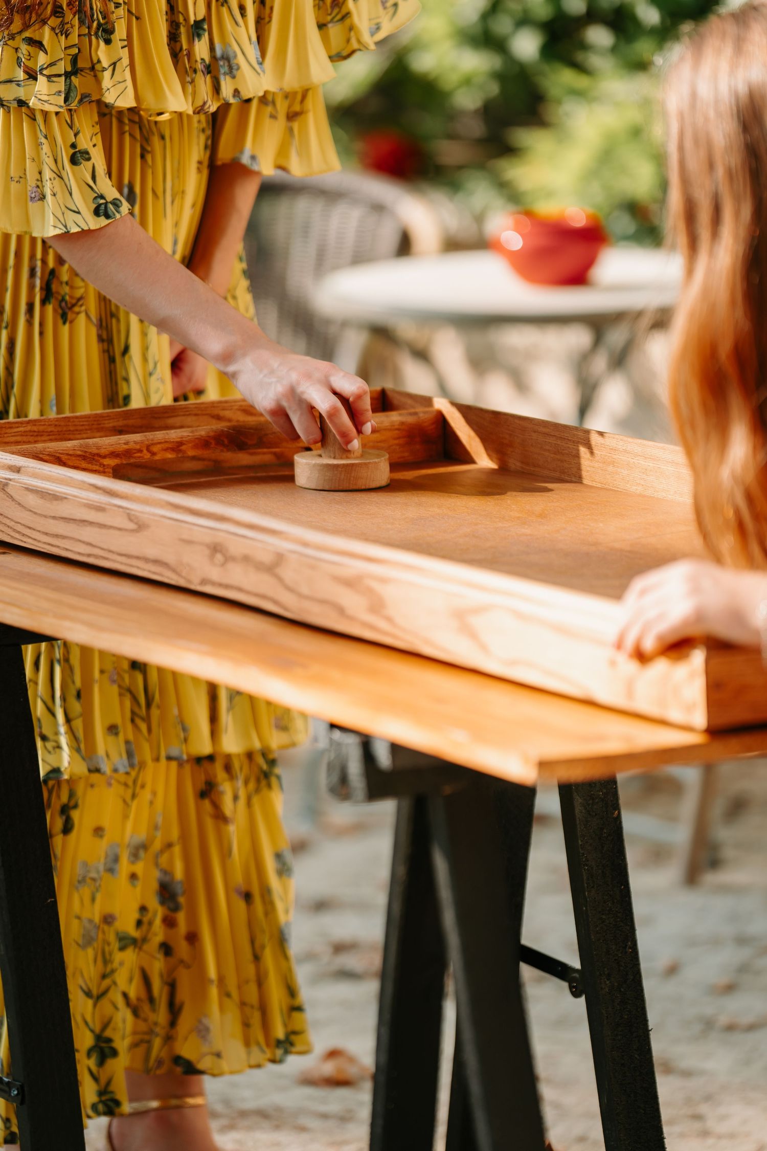Person in yellow dress sanding a wooden board on black sawhorses outdoors in natural sunlight.