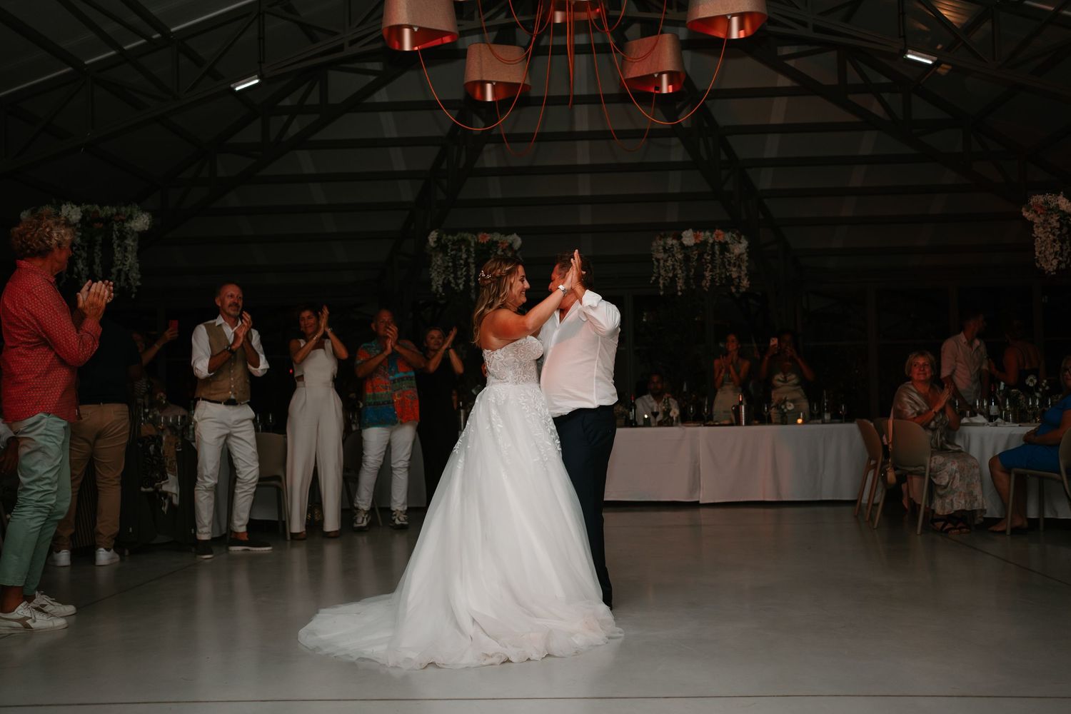 A couple performs their first dance at a wedding reception on a dimly lit dance floor with guests watching.