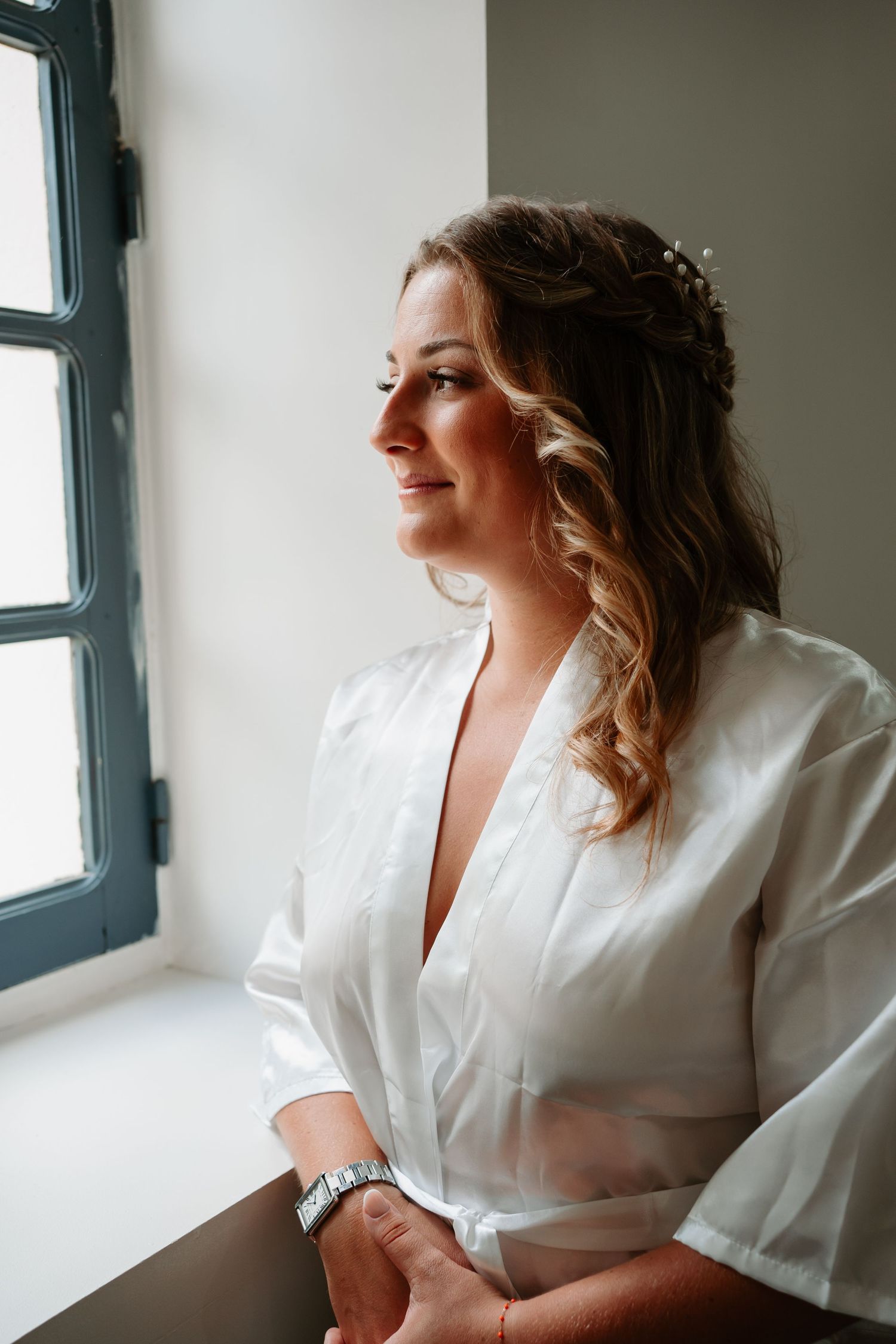 A woman in a white robe sits by a window in a series of intimate boudoir photos with soft natural lighting.