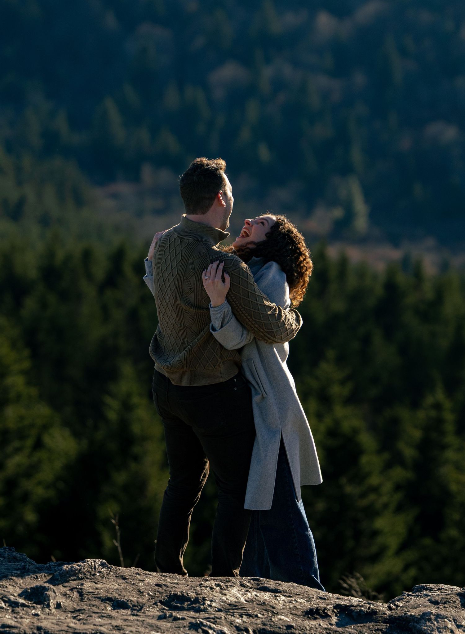 Couple embracing and laughing on a rocky mountain overlook with a sunlit evergreen forest stretching out behind them.