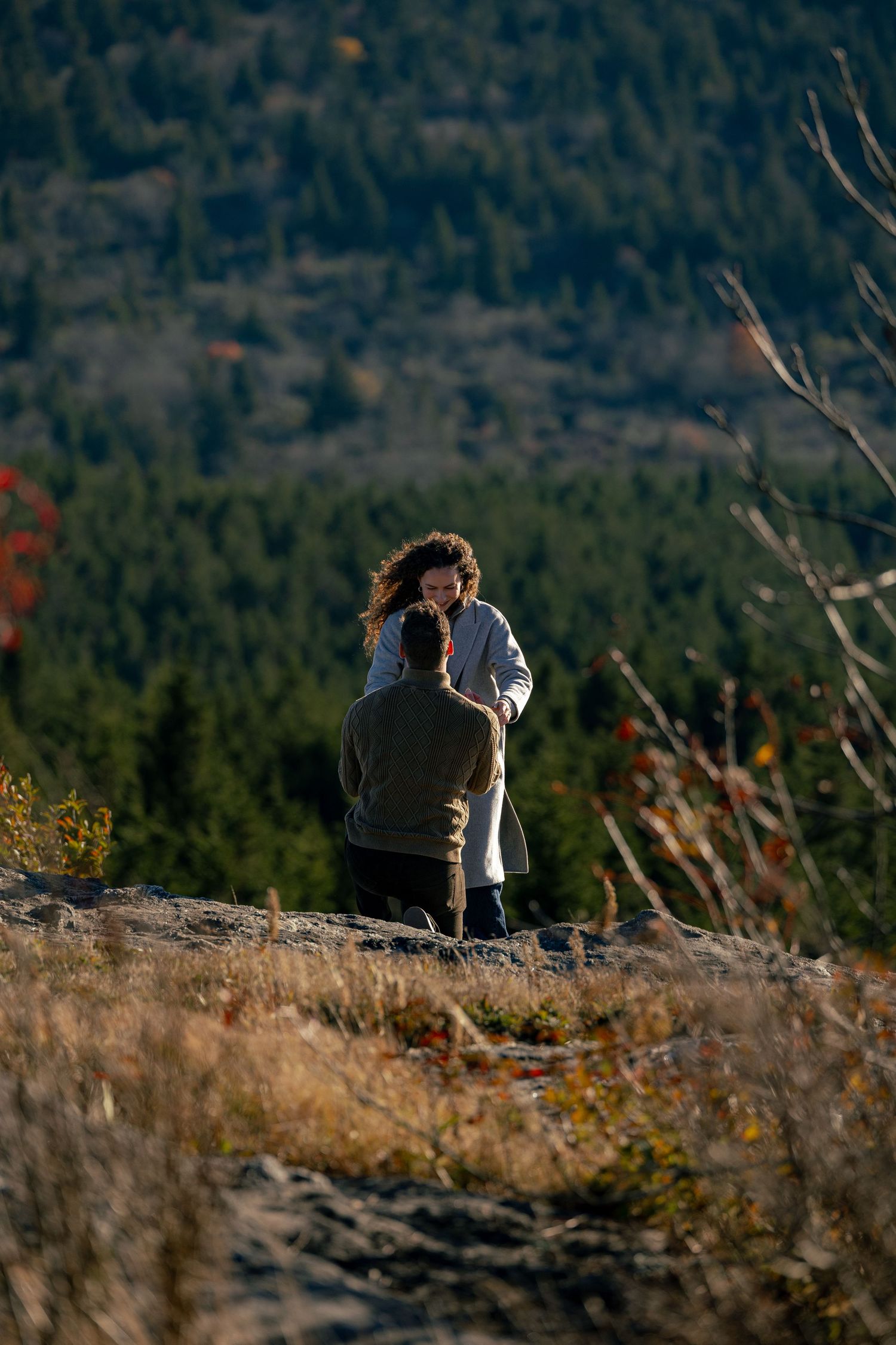 Man kneeling on a rocky mountain overlook facing a curly-haired woman, with evergreen forests and autumn foliage in the background.