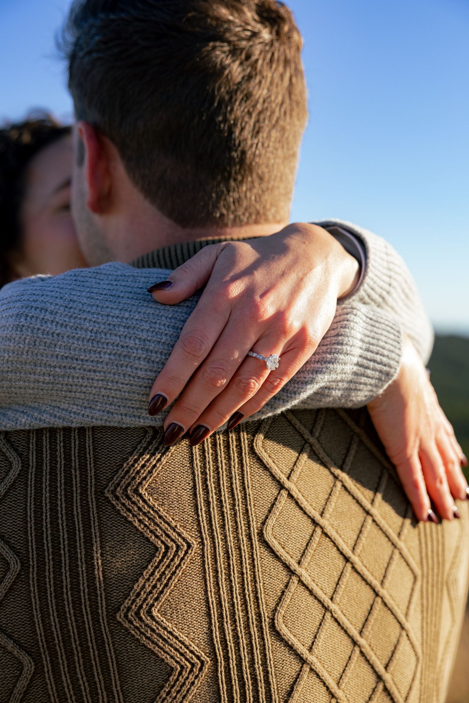 Close-up of a woman's hand with dark nail polish and a diamond engagement ring resting on her partner's shoulder in a warm embrace.