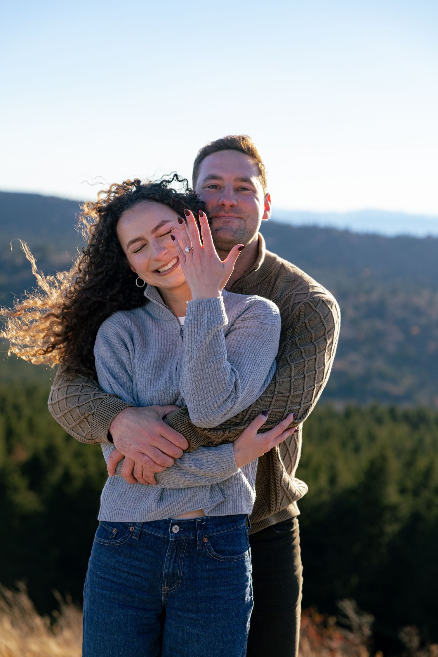 Smiling woman with curly hair shows her engagement ring as her fiancé hugs her from behind on a scenic mountain overlook.